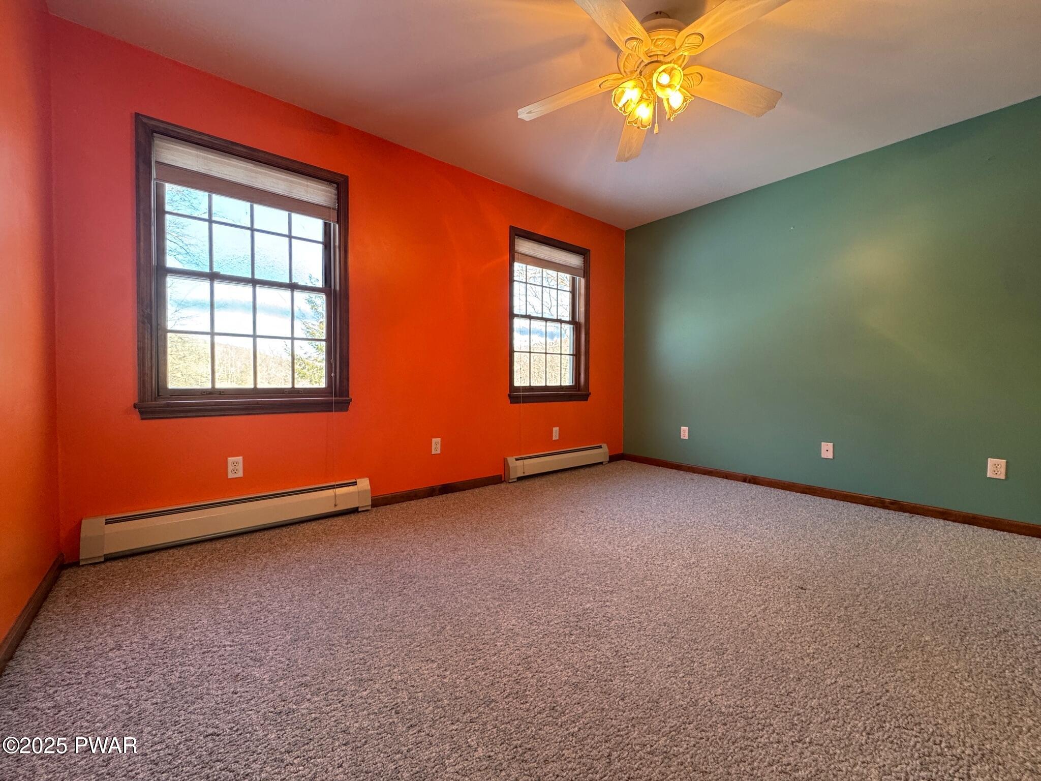 Empty bedroom with bright orange and green painted walls, two windows, carpeted floor, and a ceiling fan light.