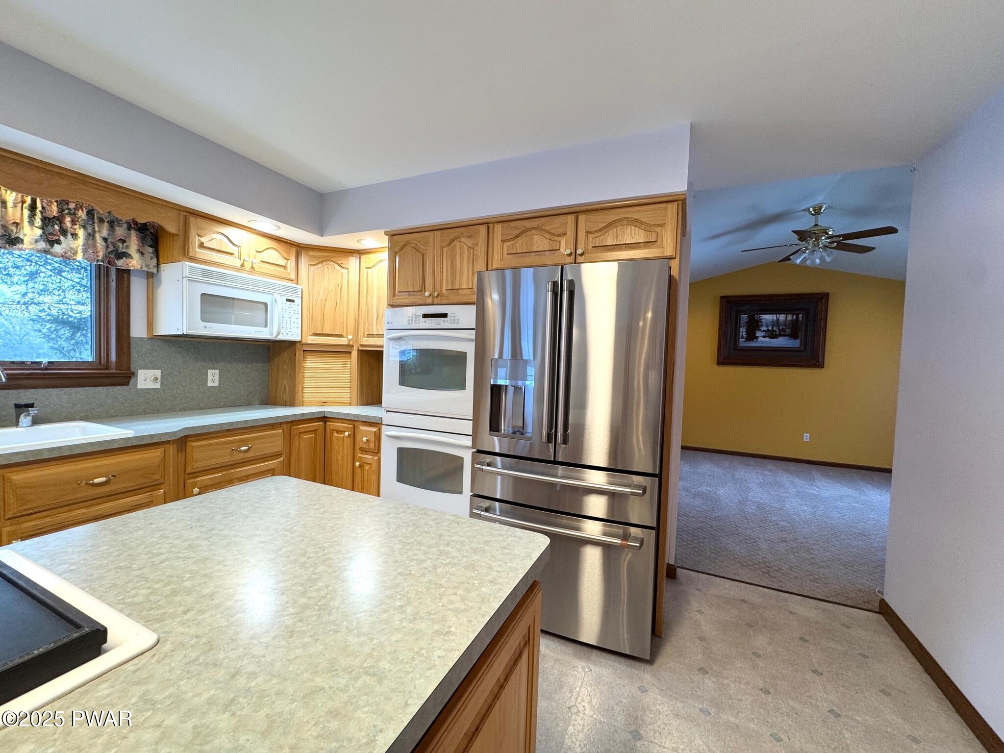 Kitchen with oak cabinets, stainless steel refrigerator, white wall oven and microwave, laminate countertops, and a view into an adjacent carpeted room with a ceiling fan.