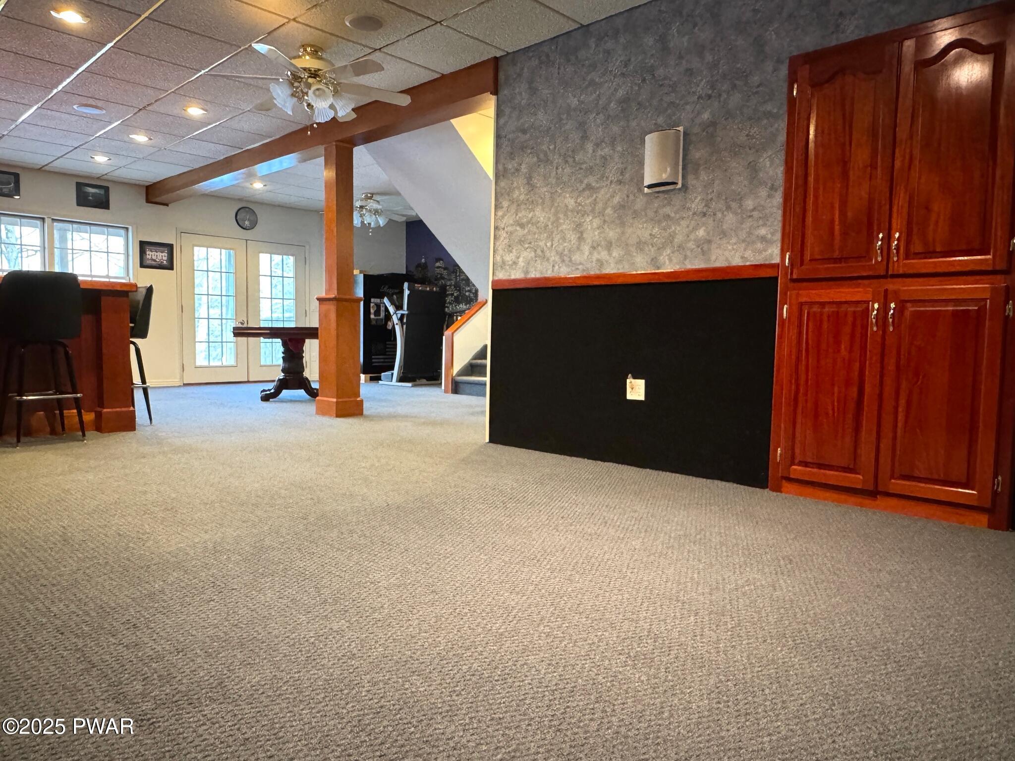 Finished basement recreation room with carpet, built-in wood cabinets, bar area, French doors, and a staircase in the background.