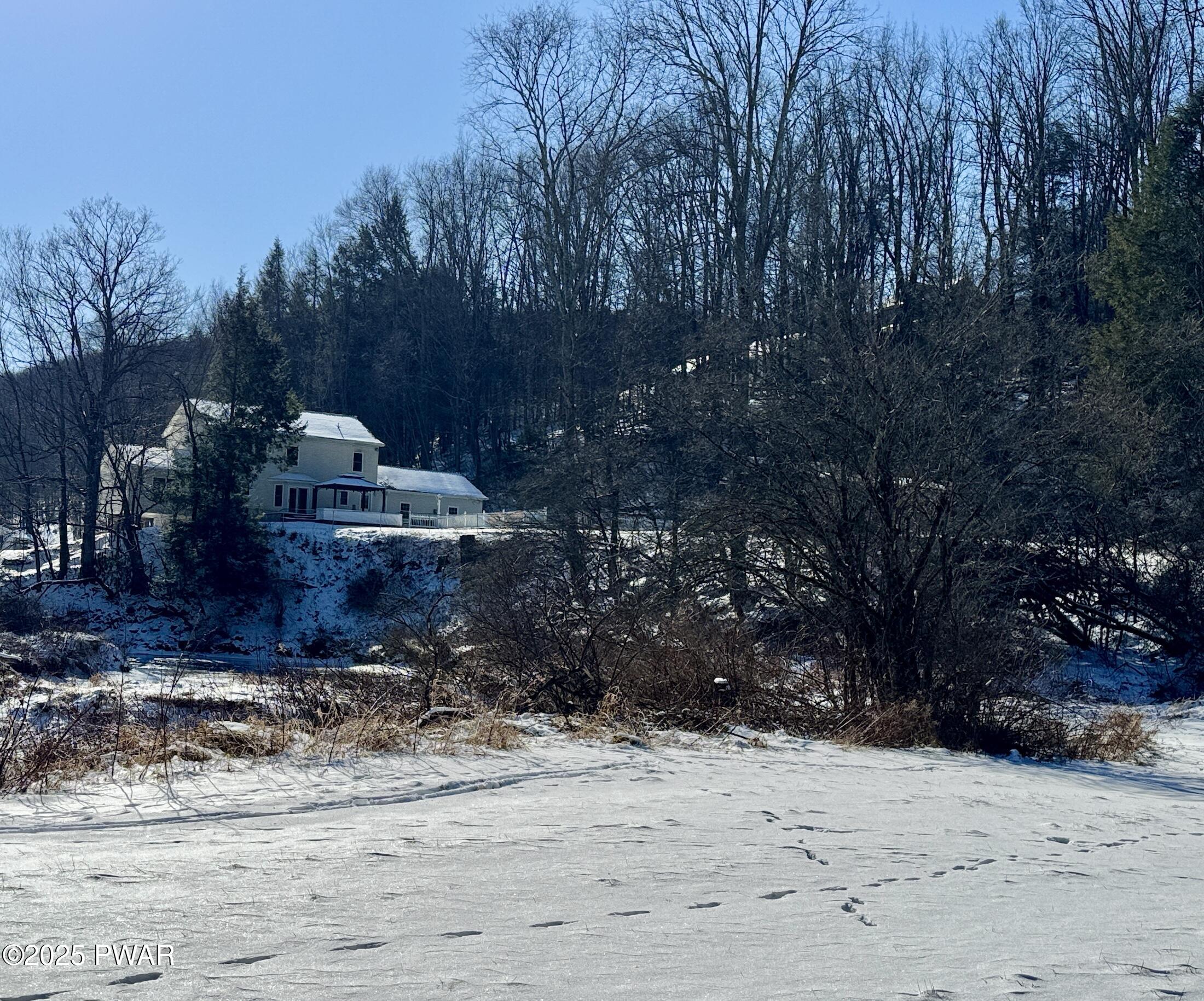 Snow-covered yard or lot with a house set back among trees on a winter day.