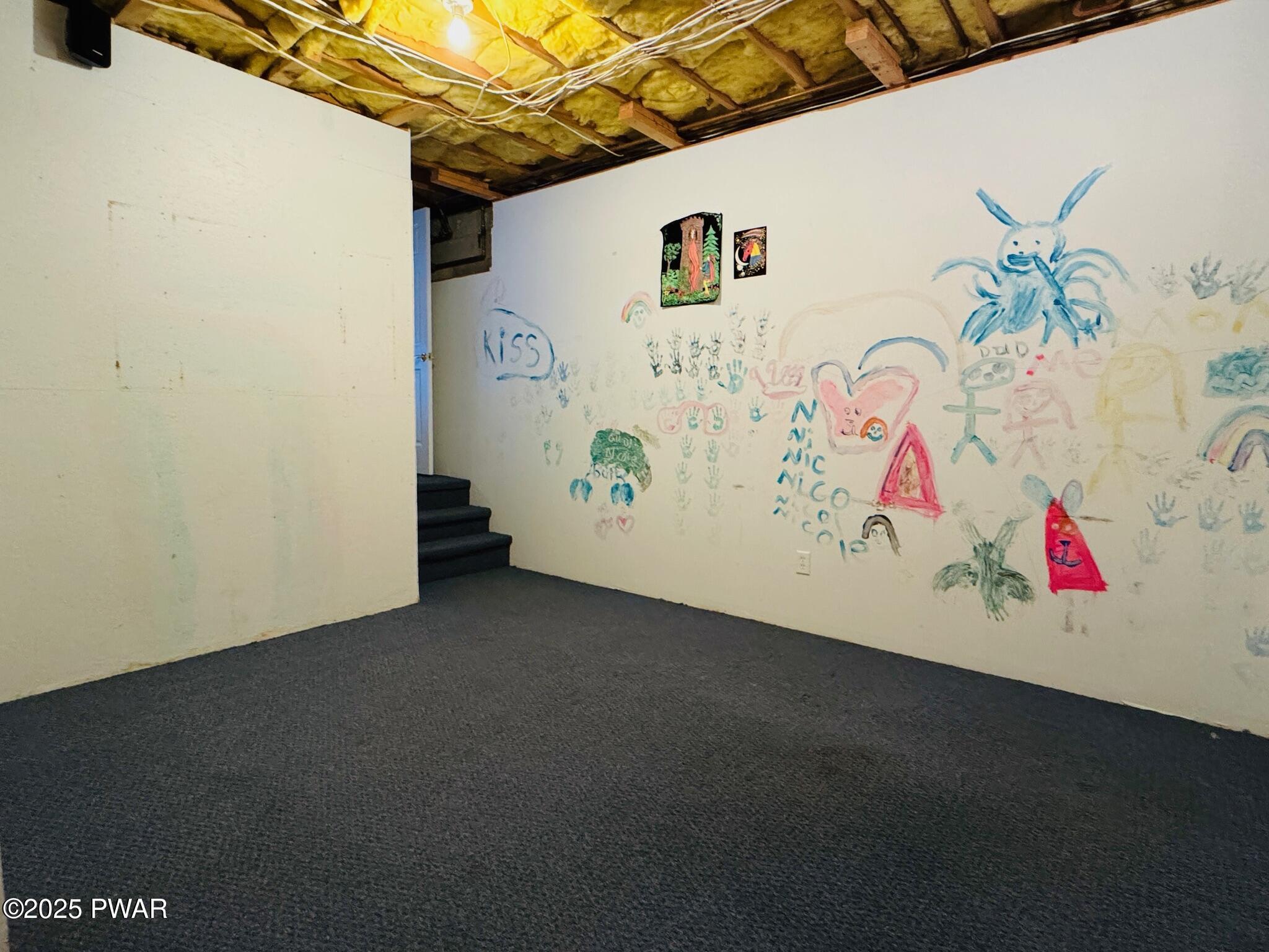 Basement room with exposed ceiling insulation, painted walls covered in children’s drawings, carpeted floor, and a small stair opening.