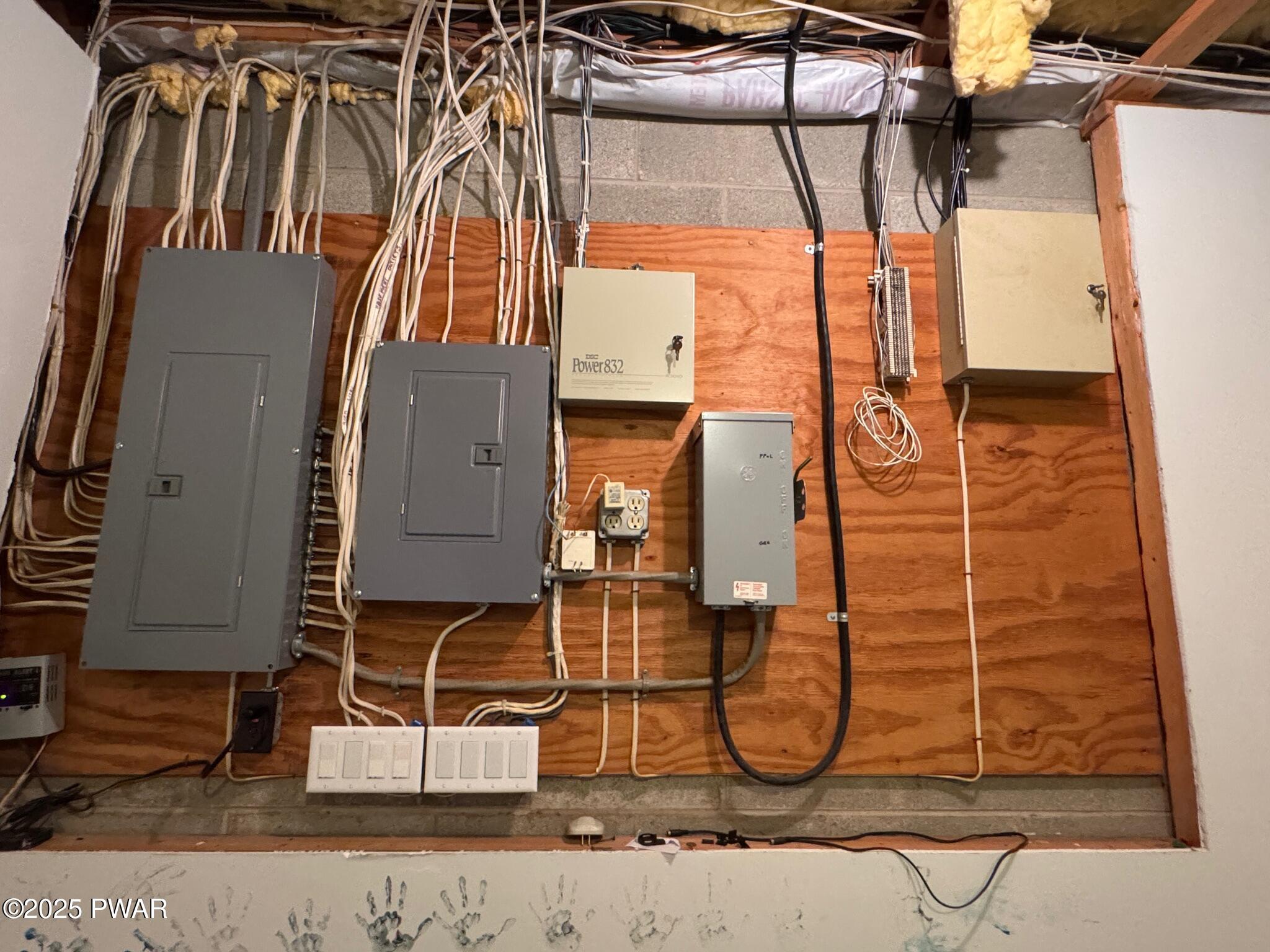 A utility room wall with multiple electrical panels, conduit, exposed wiring, and switches mounted on plywood backing.