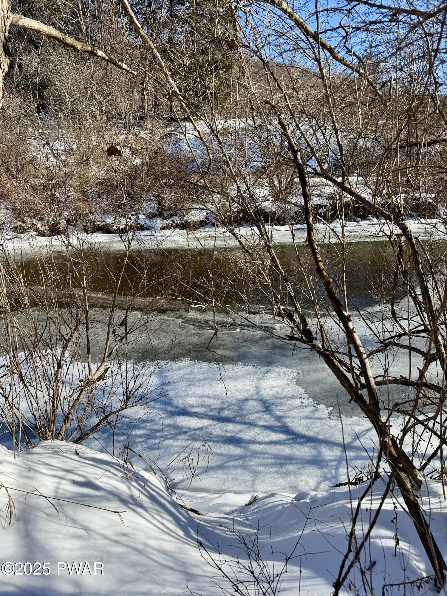 Snow-covered riverbank with leafless trees and a partially frozen river or stream in winter.