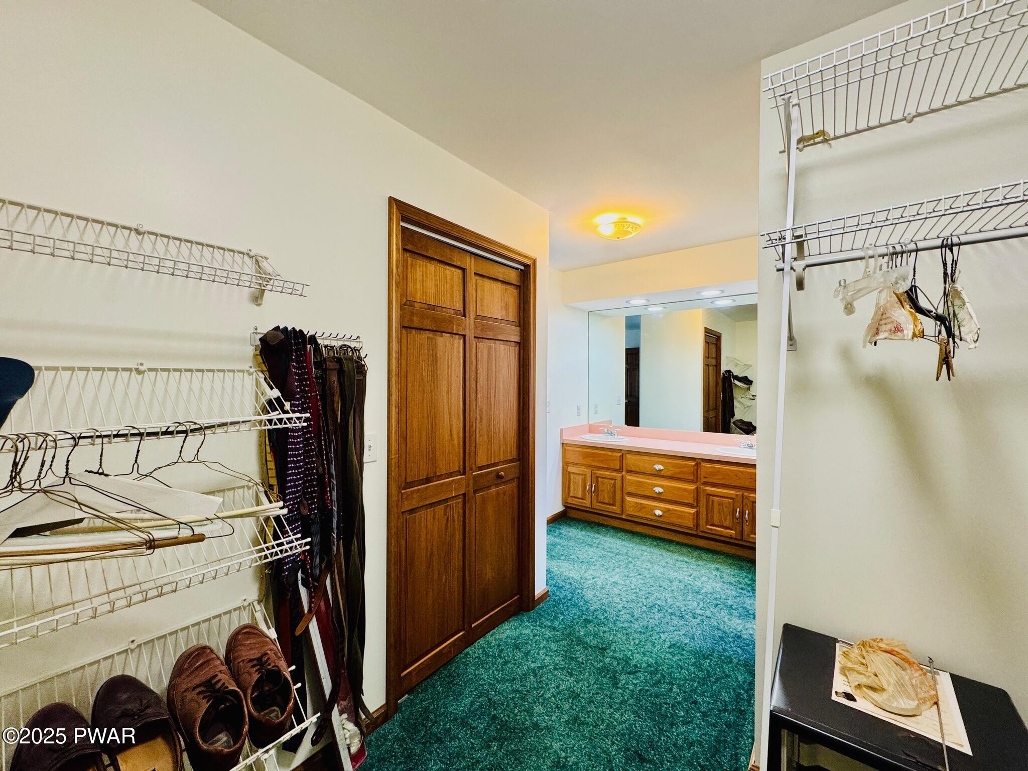 Walk-in closet with wire shelving, hanging clothes and shoes, opening into a bathroom vanity area with carpet and wood doors.