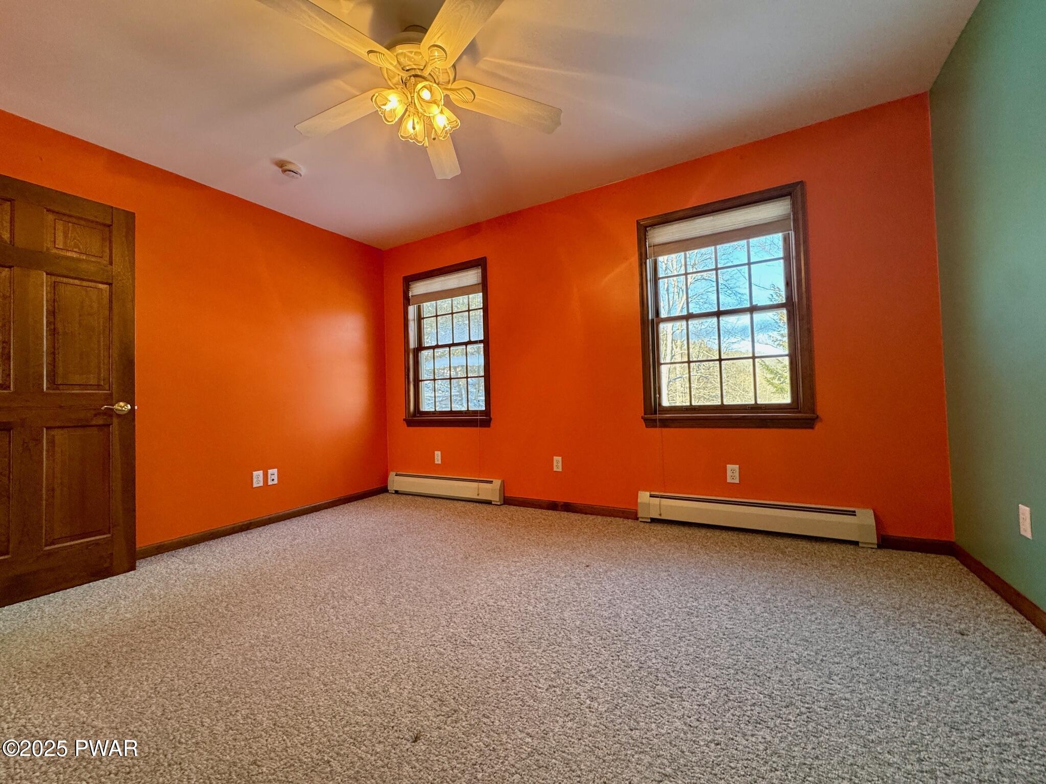 Empty bedroom with orange walls, carpet, two windows, a ceiling fan, and a wooden door.