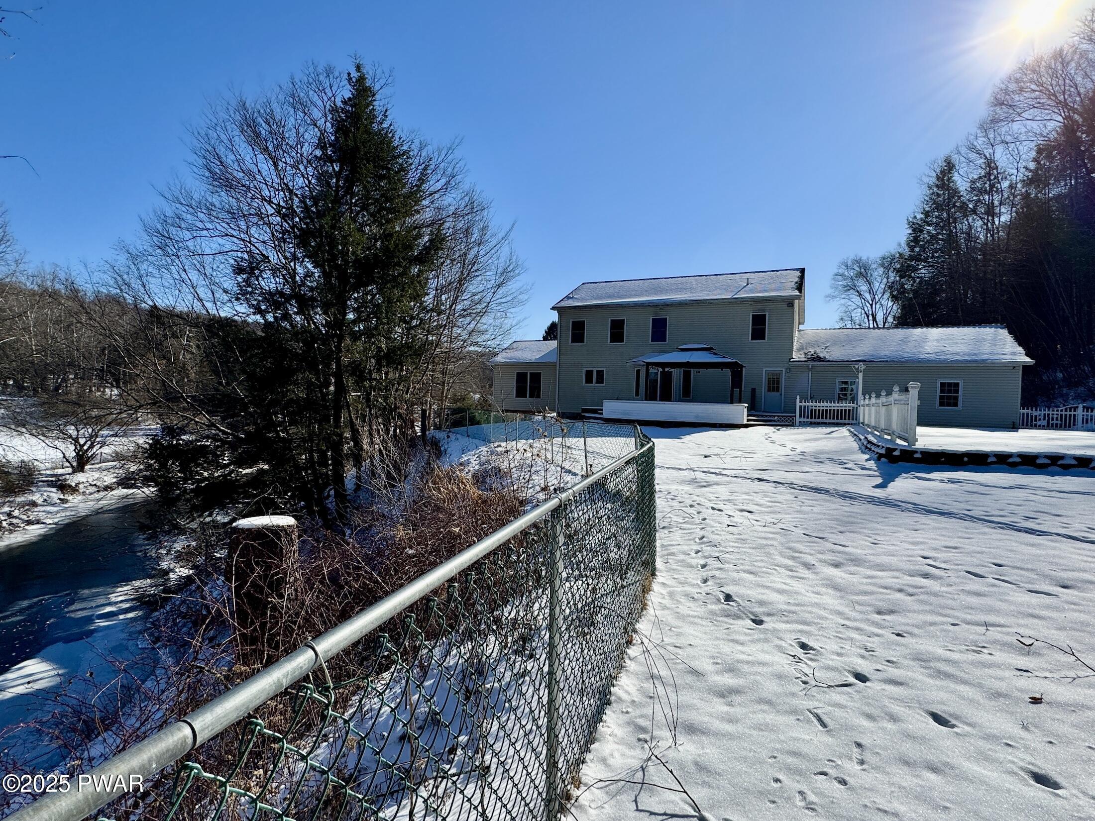 Snow-covered backyard with a fenced path, trees, a small stream, and the rear of a two-story house with a deck and patio area under a bright blue sky.