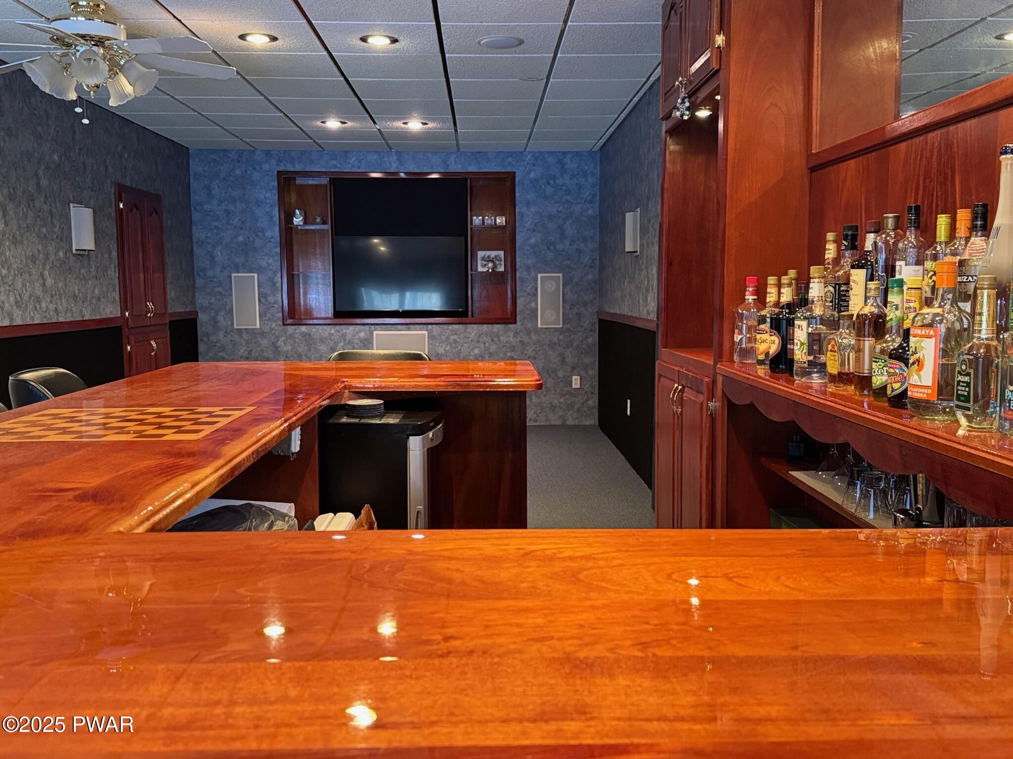 A finished basement entertainment room with a glossy wood bar, built-in shelving stocked with liquor bottles, and a wall-mounted TV.