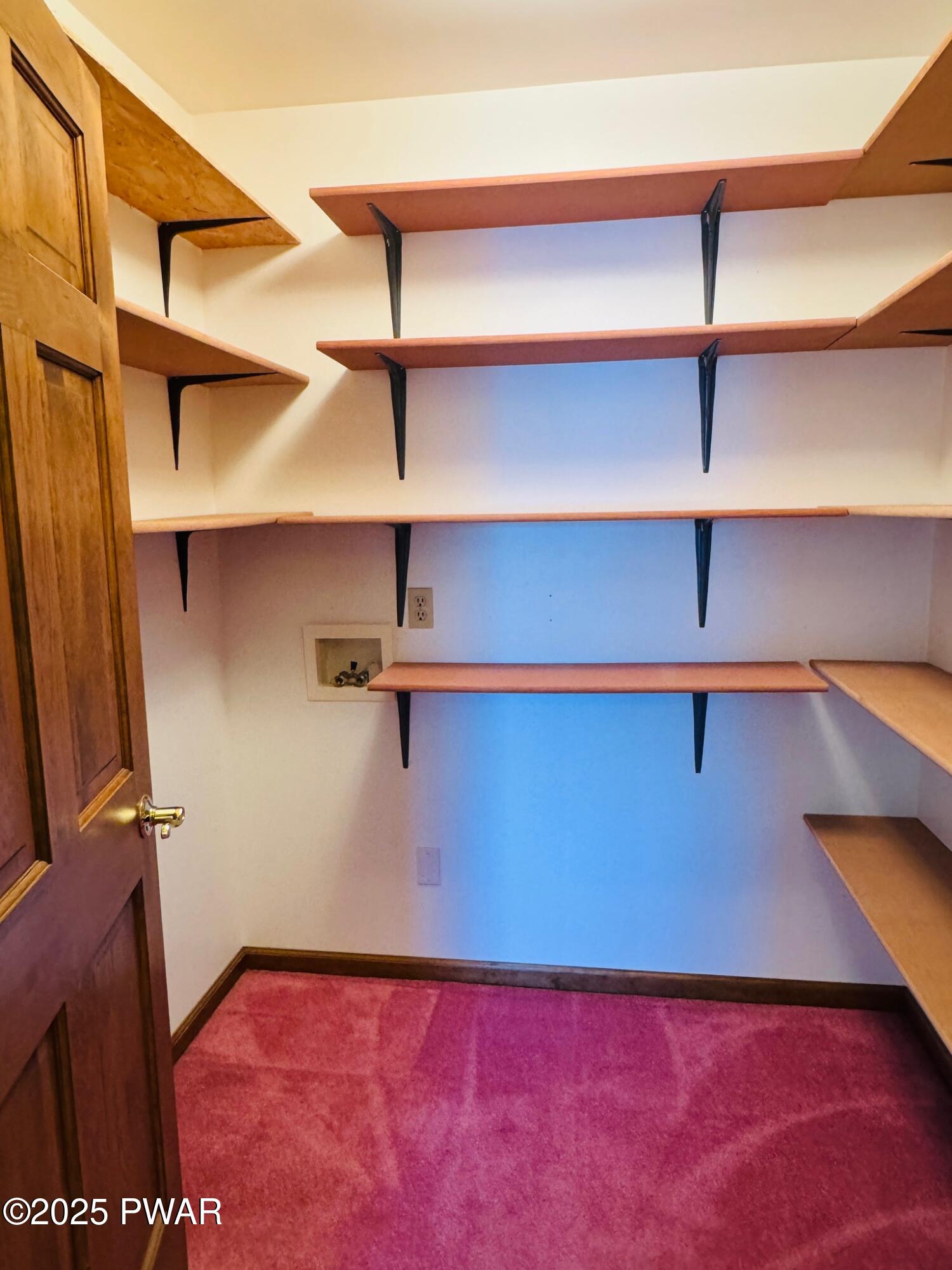 Walk-in closet with multiple wooden shelves and pink carpet flooring, viewed through an open door.