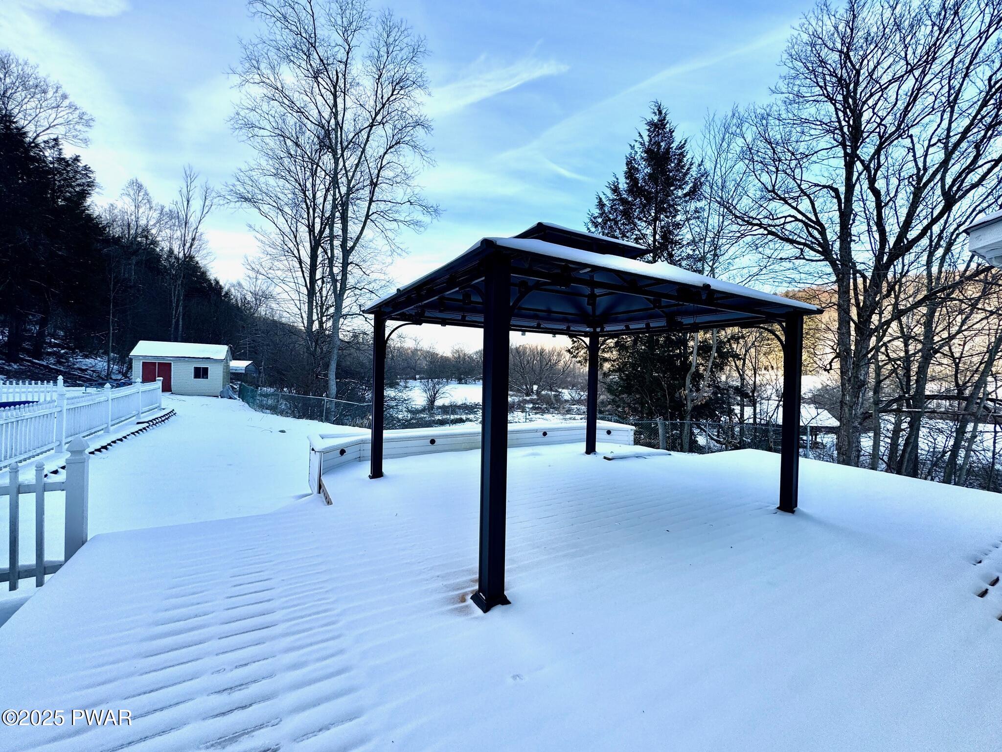 Snow-covered backyard patio or deck with a black gazebo, white fence, and trees in winter.