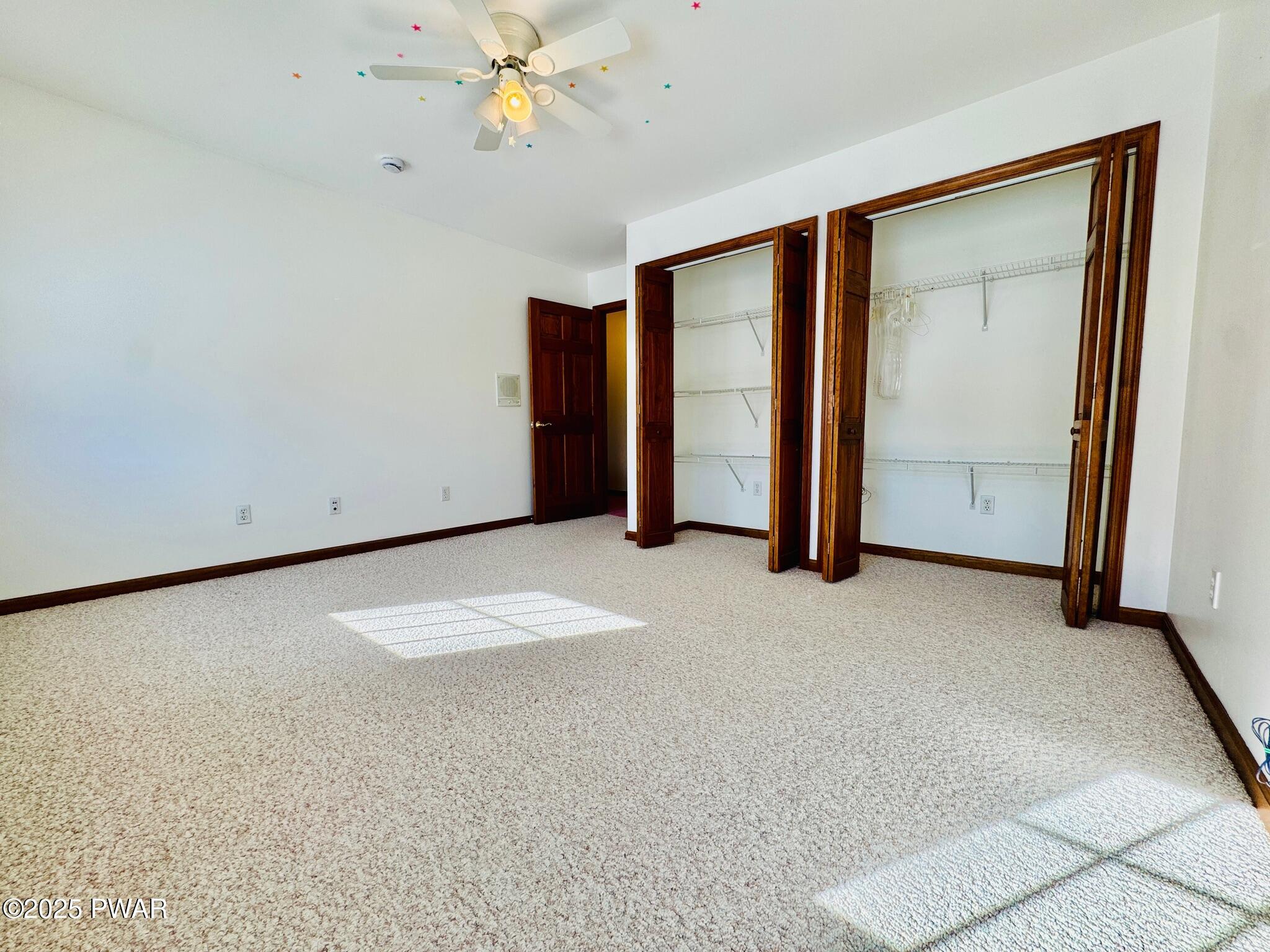Empty carpeted bedroom with white walls, ceiling fan, and two open closet areas with wire shelving.