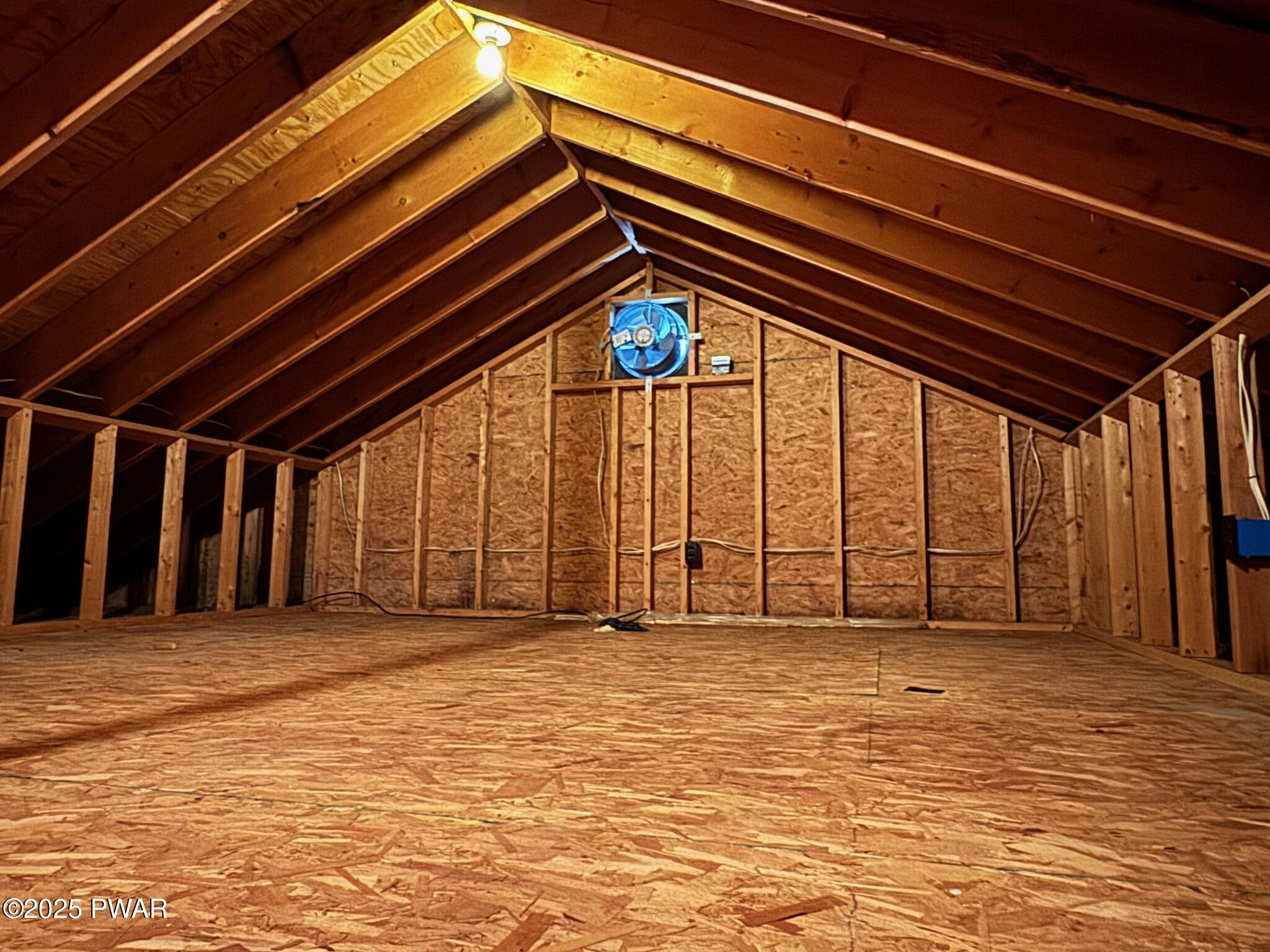 Unfinished attic space with exposed wooden rafters, insulation, and a small fan on the far wall.