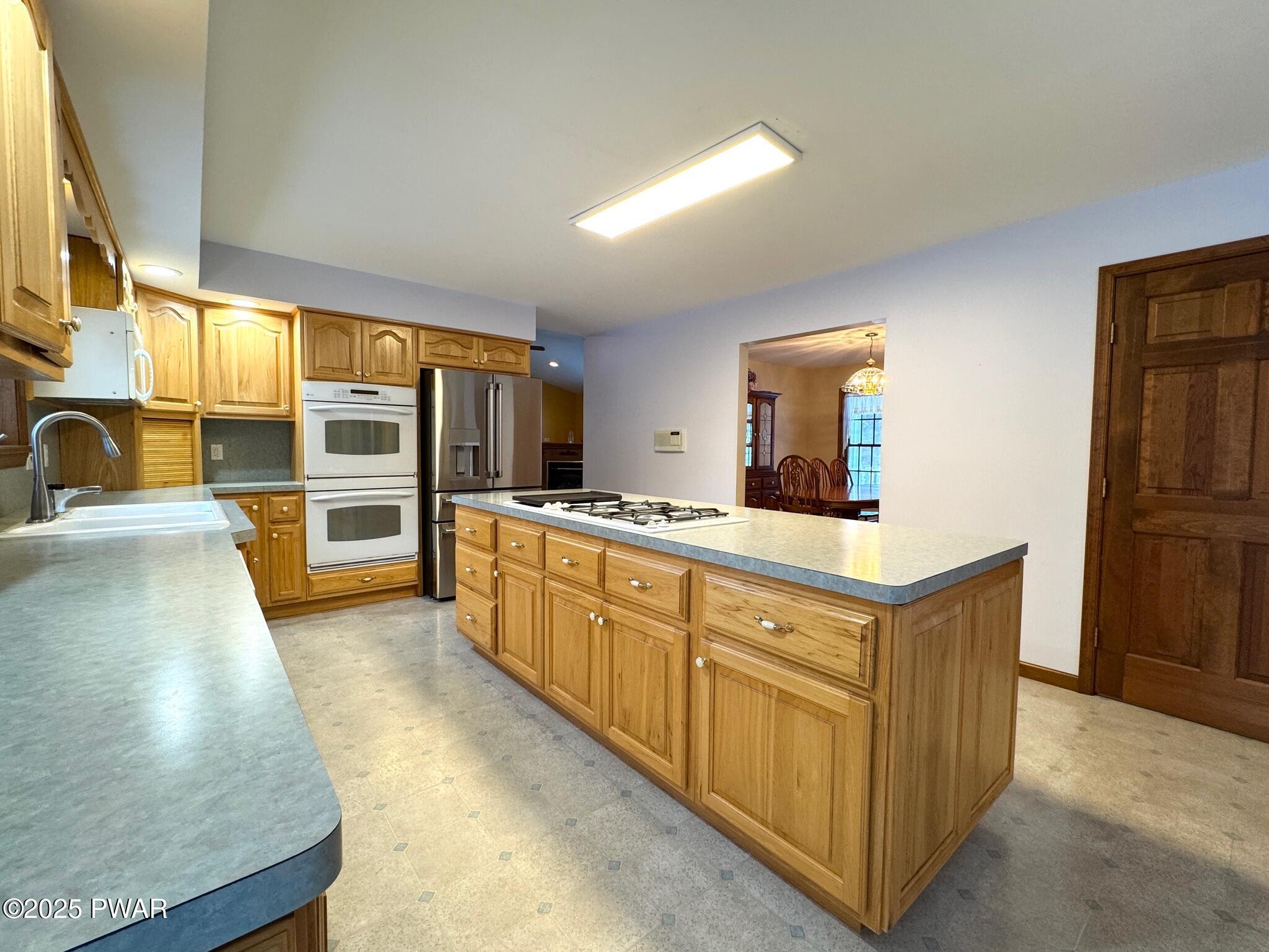 Spacious kitchen with wooden cabinets, center island with cooktop, stainless refrigerator, built-in oven, and view into adjacent dining area.