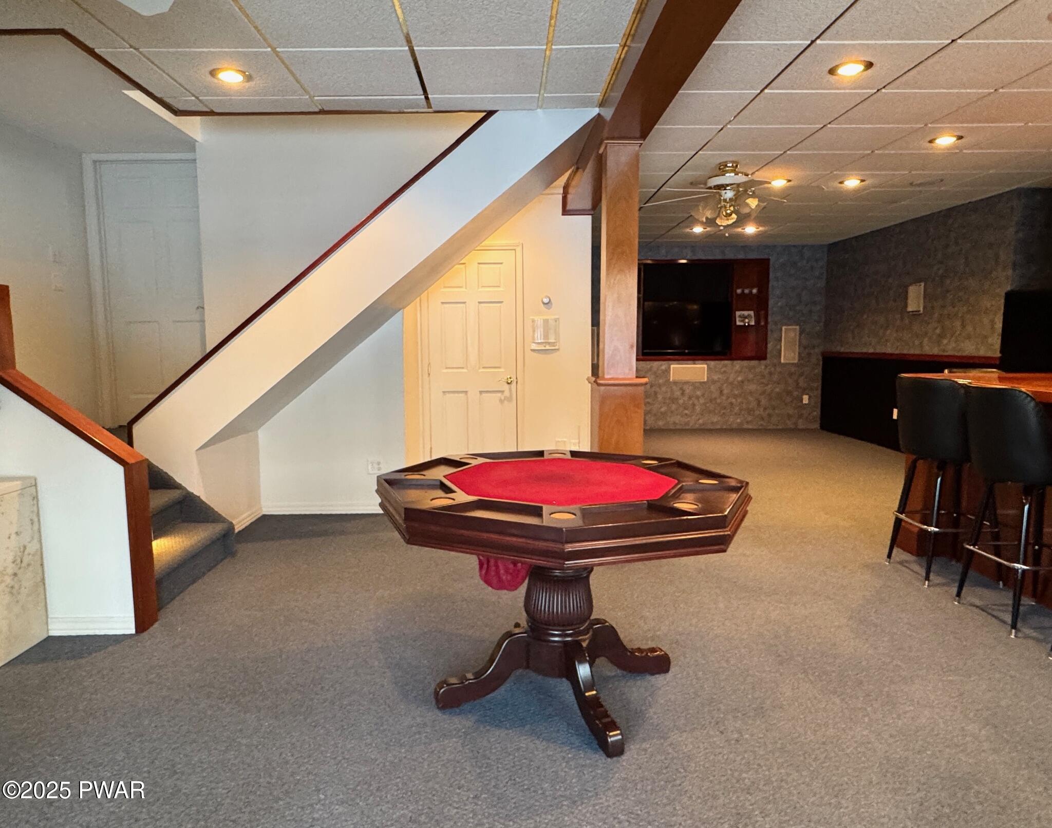 Finished basement recreation room with a poker table in the foreground, staircase to the left, and a bar area with seating to the right.