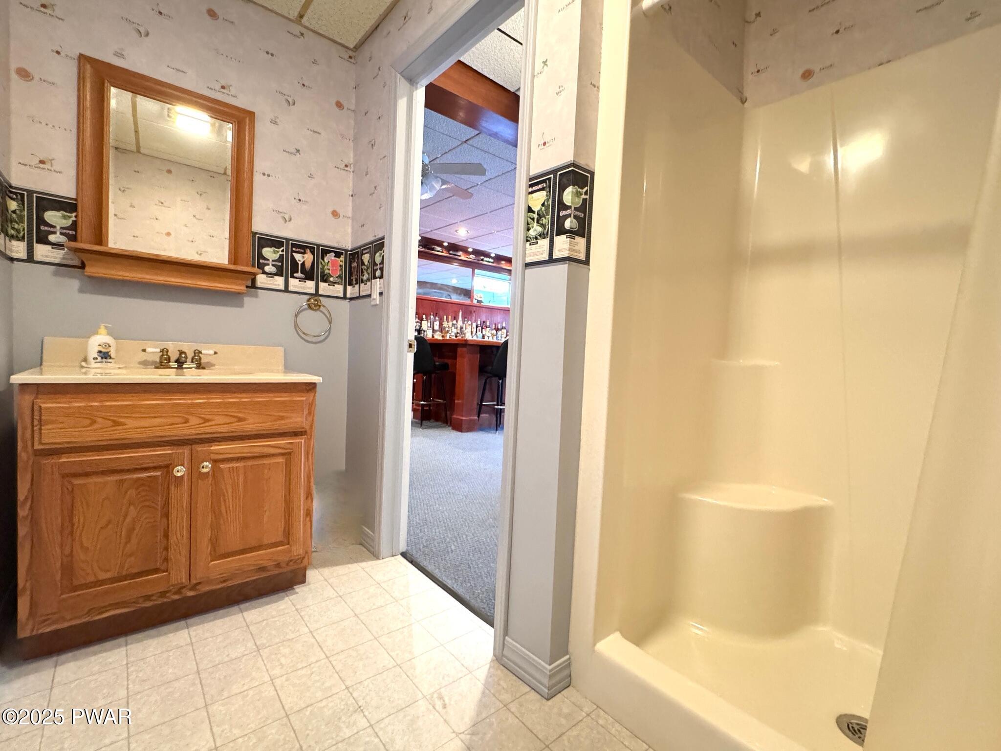 Bathroom with wood vanity, sink, mirror, and shower stall; doorway opens to a bar area beyond.
