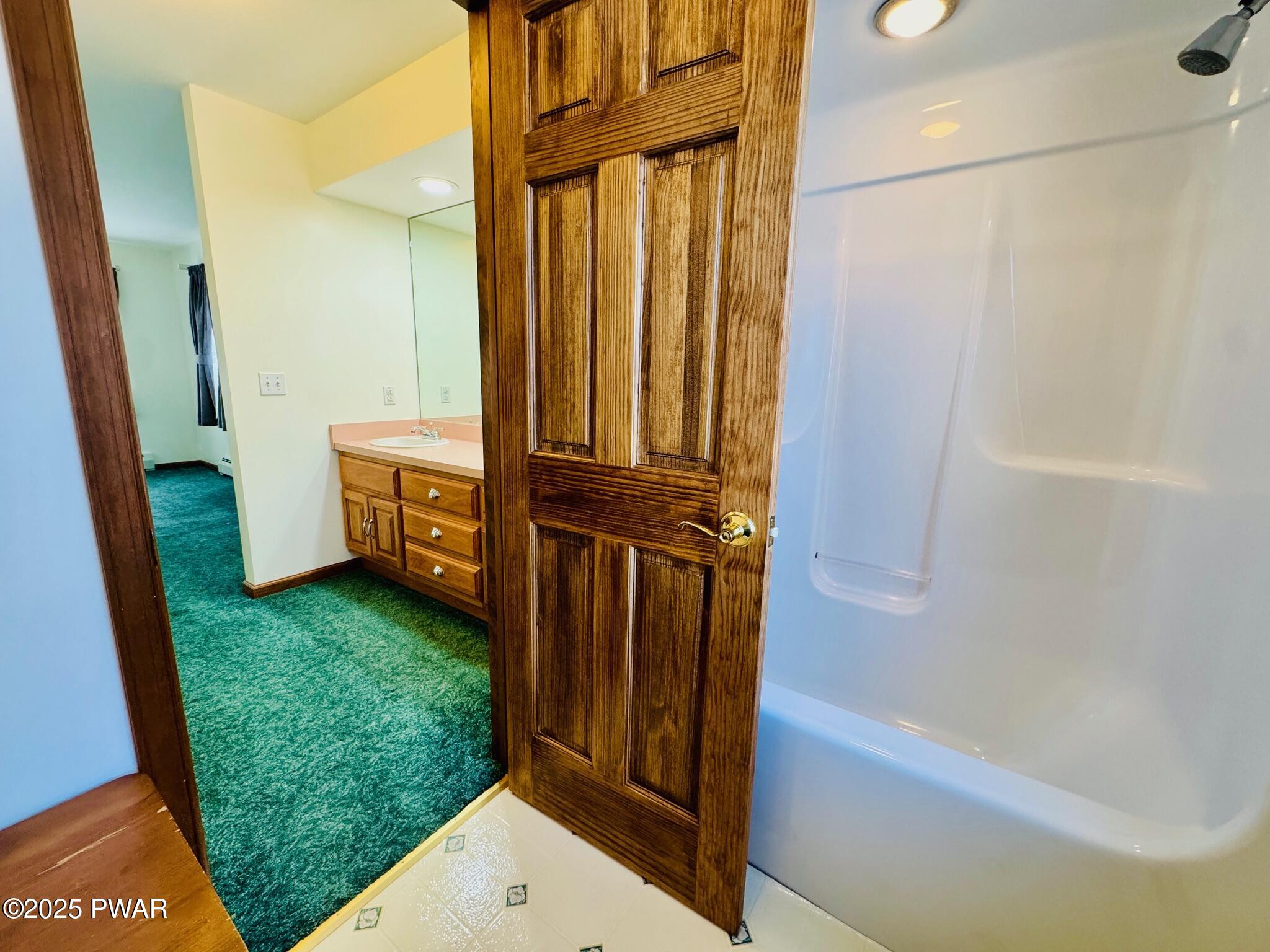 Bathroom with a white tub and shower surround, wood door, vanity with pink countertop, and green carpet visible in the adjoining room.