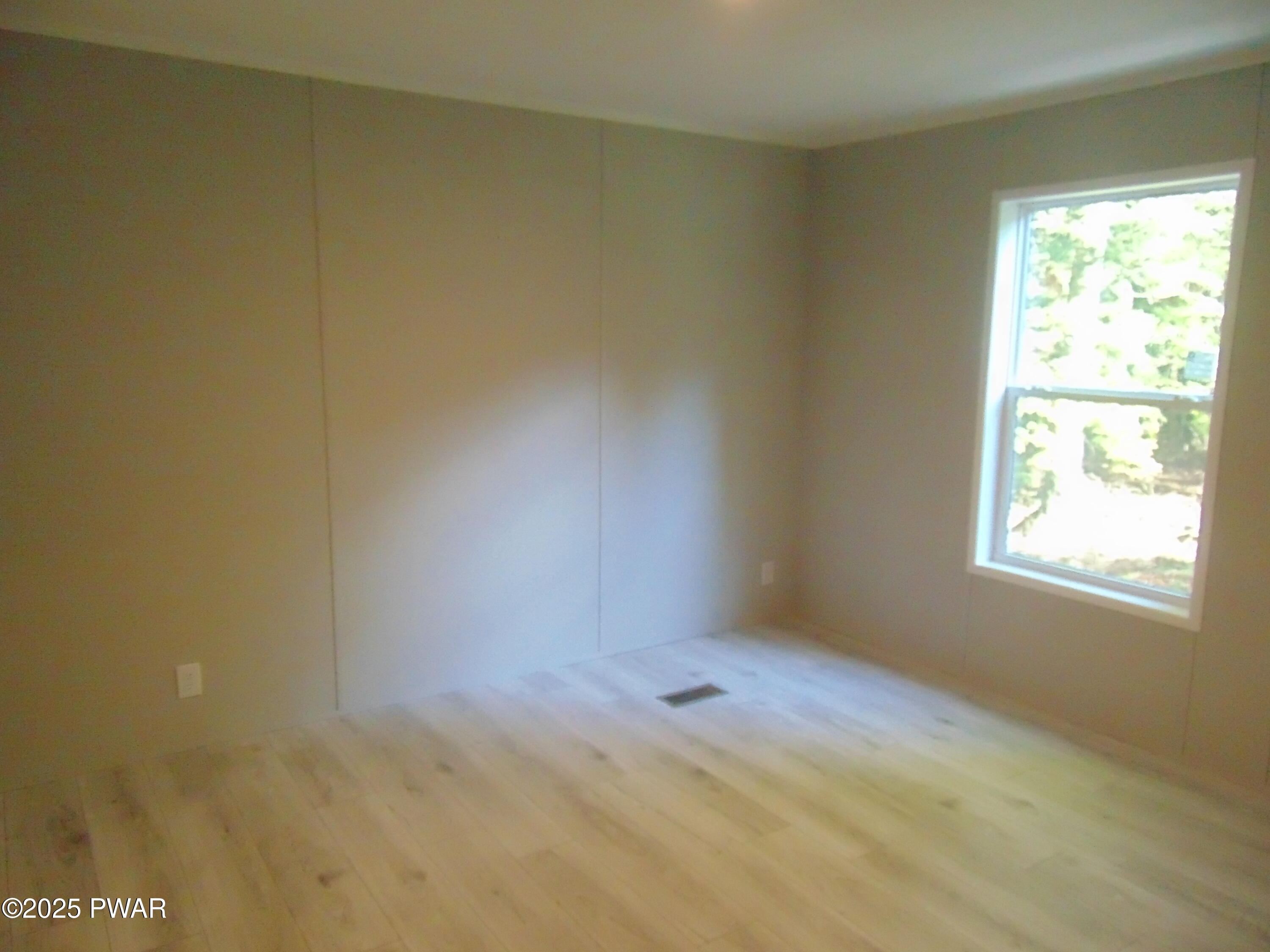 Empty bedroom with light wood floors, beige walls, and a single window letting in daylight.