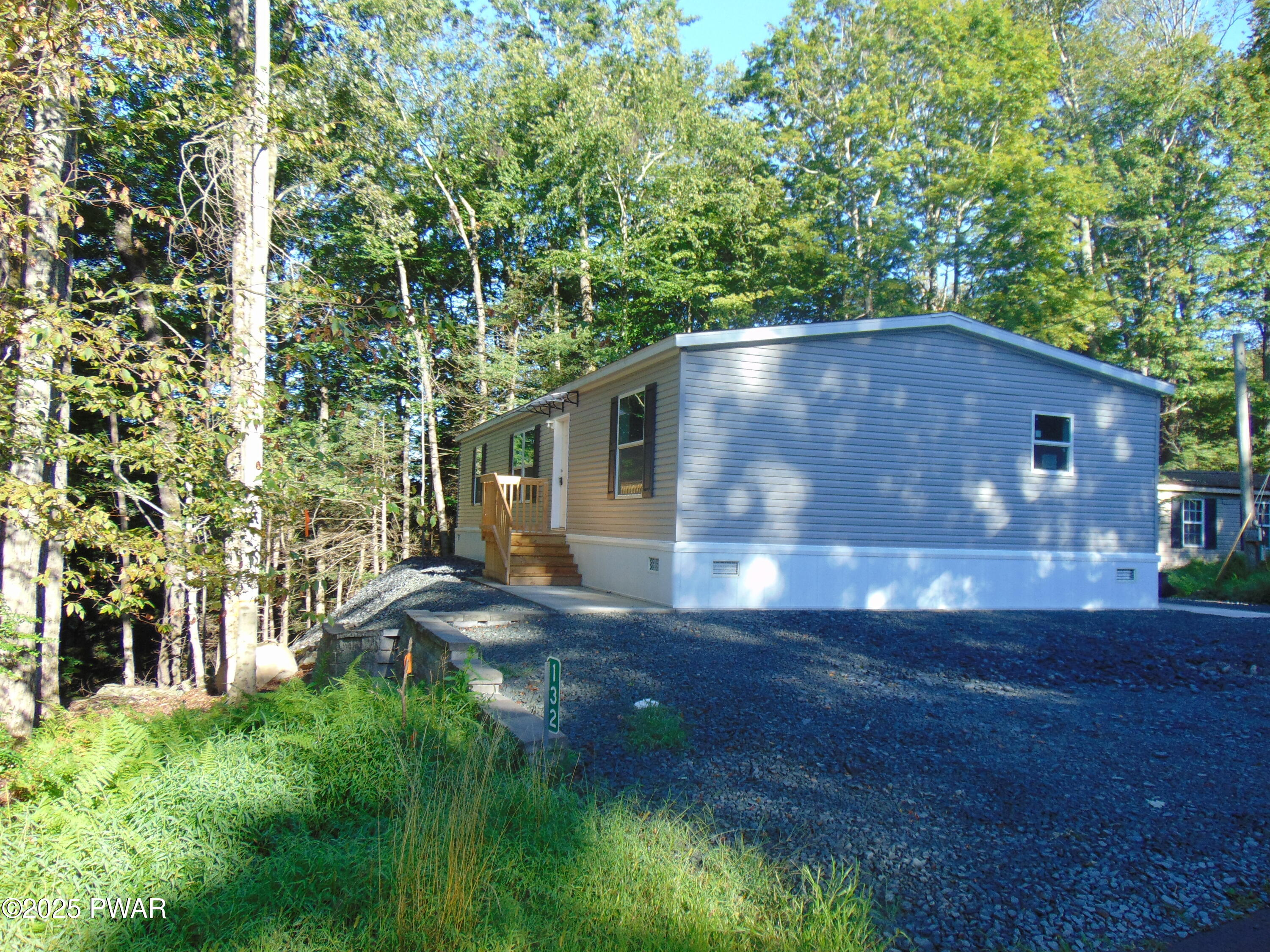 Front exterior of a gray manufactured home with a small porch and gravel driveway surrounded by trees.