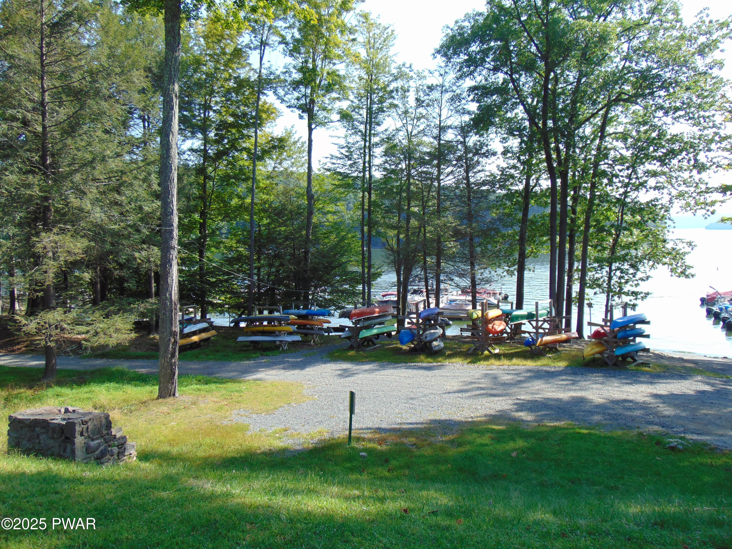 Wooded lakeside yard with racks of colorful canoes and kayaks near the water, gravel path, grass, and a dock visible through the trees.