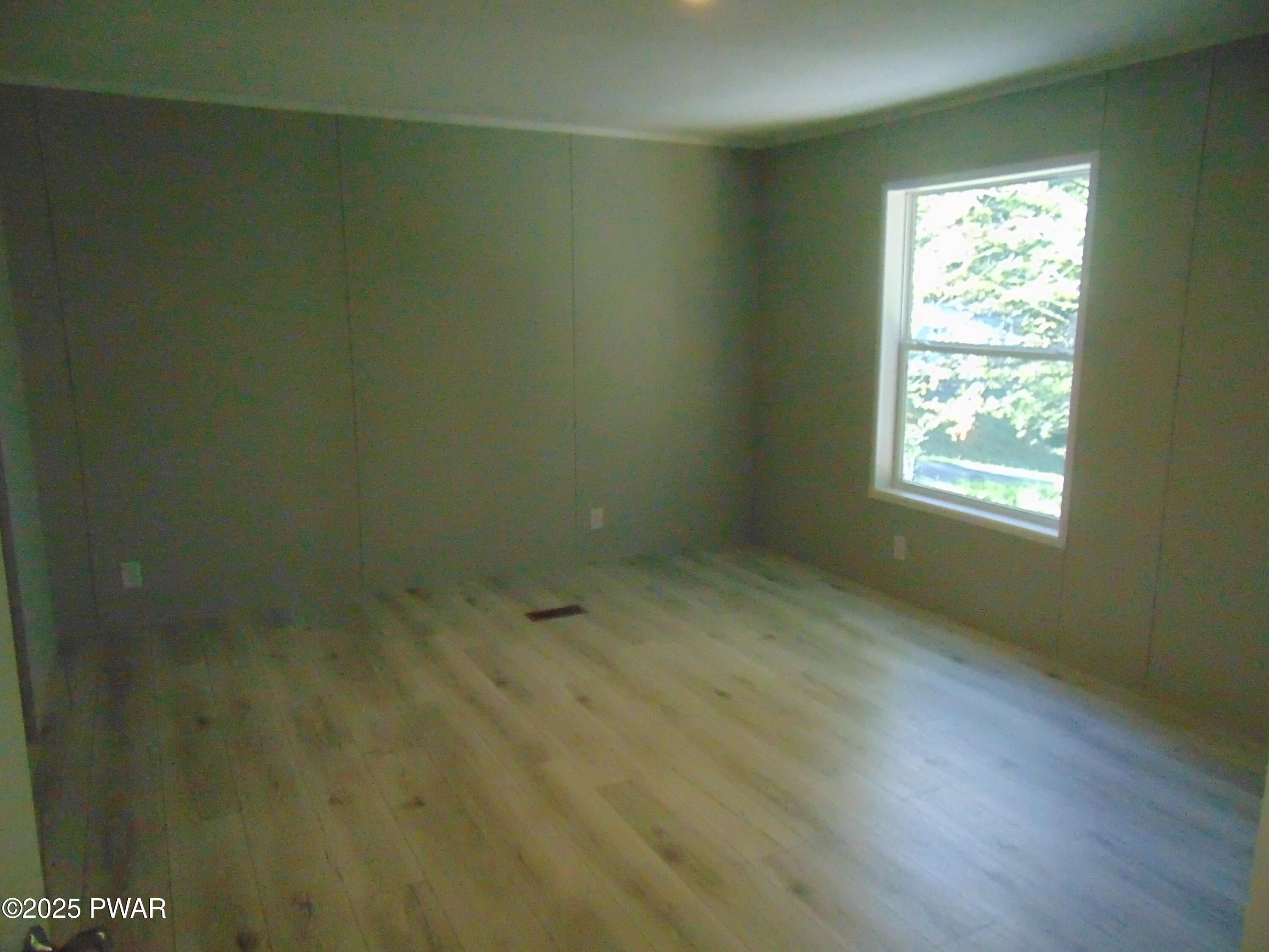 An empty bedroom with wood floors, tan walls, and one window letting in daylight.