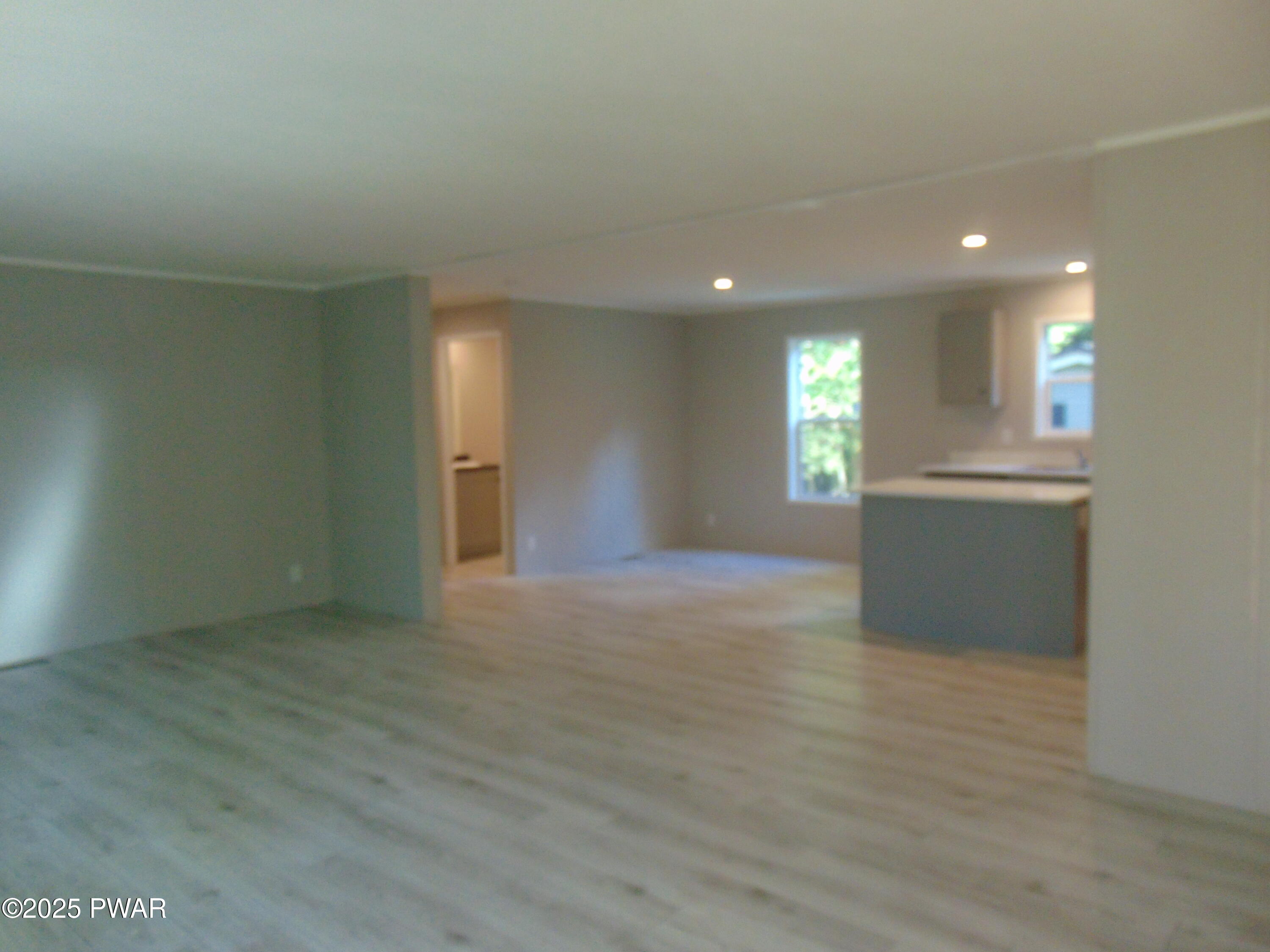 Empty open-plan living room with light wood floors, recessed lighting, and a view into the kitchen area with windows.