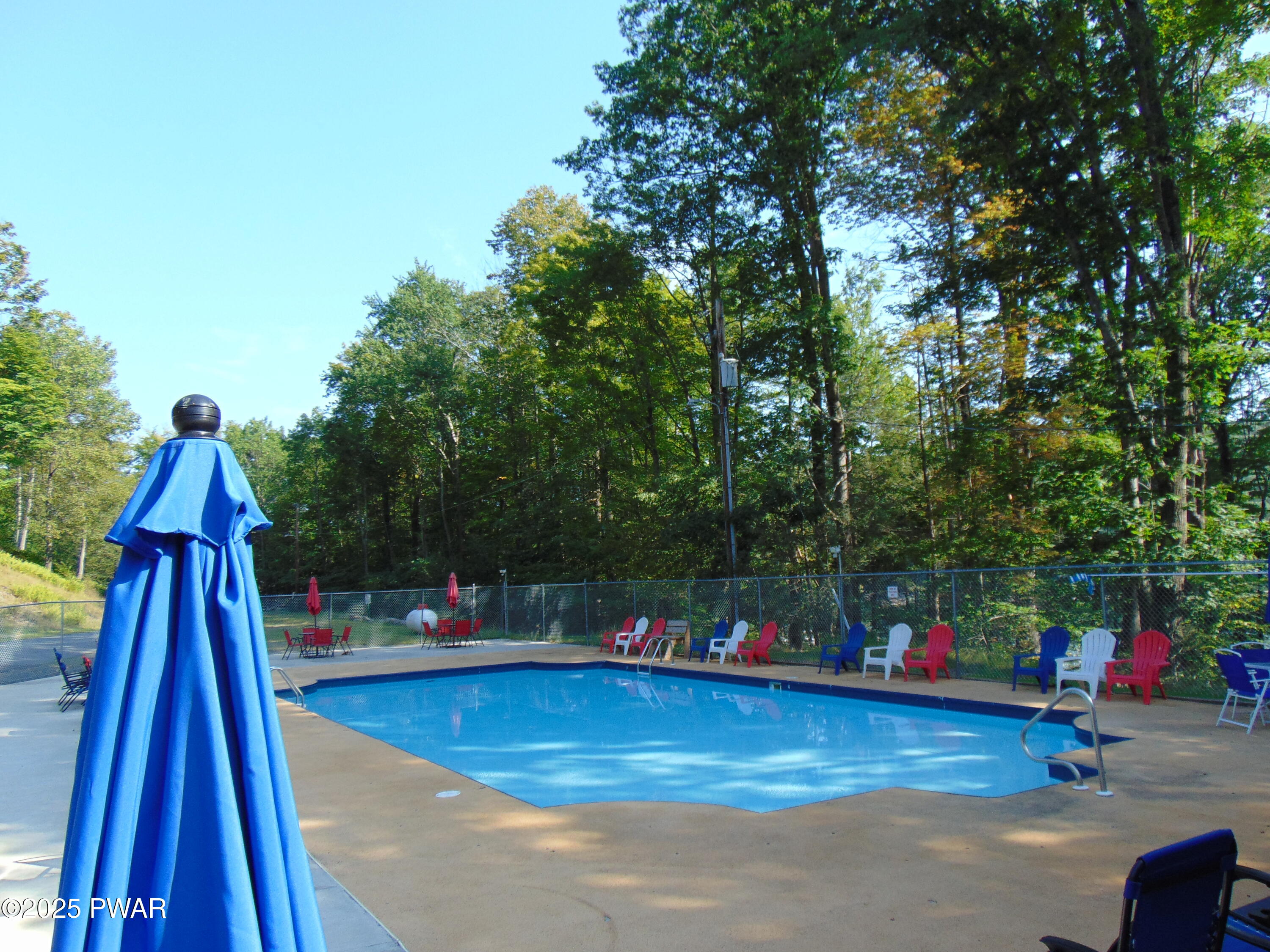 Outdoor swimming pool surrounded by chairs, tables, and umbrellas with trees in the background.