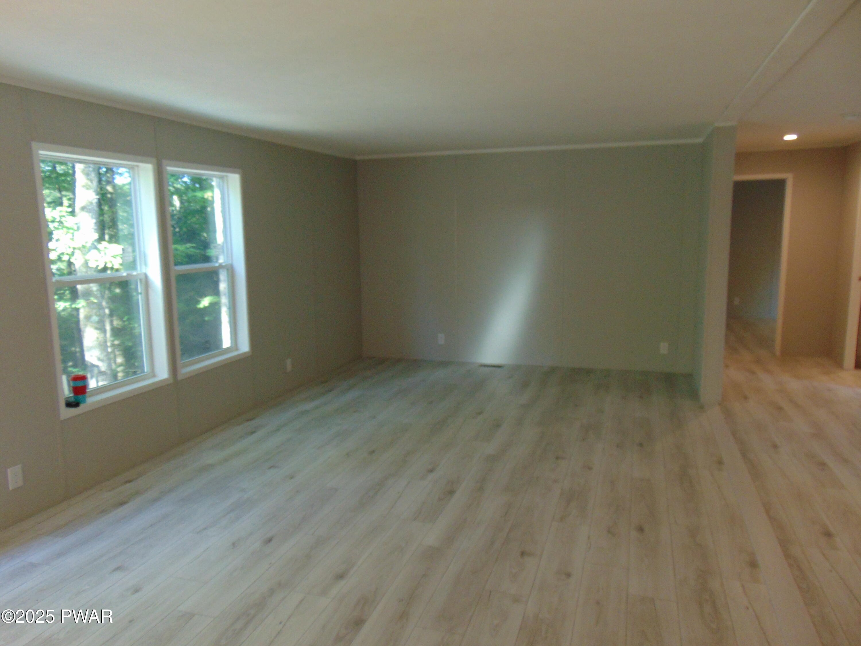 Empty living room with light wood floors, gray walls, two large windows, and a doorway to an adjoining area.