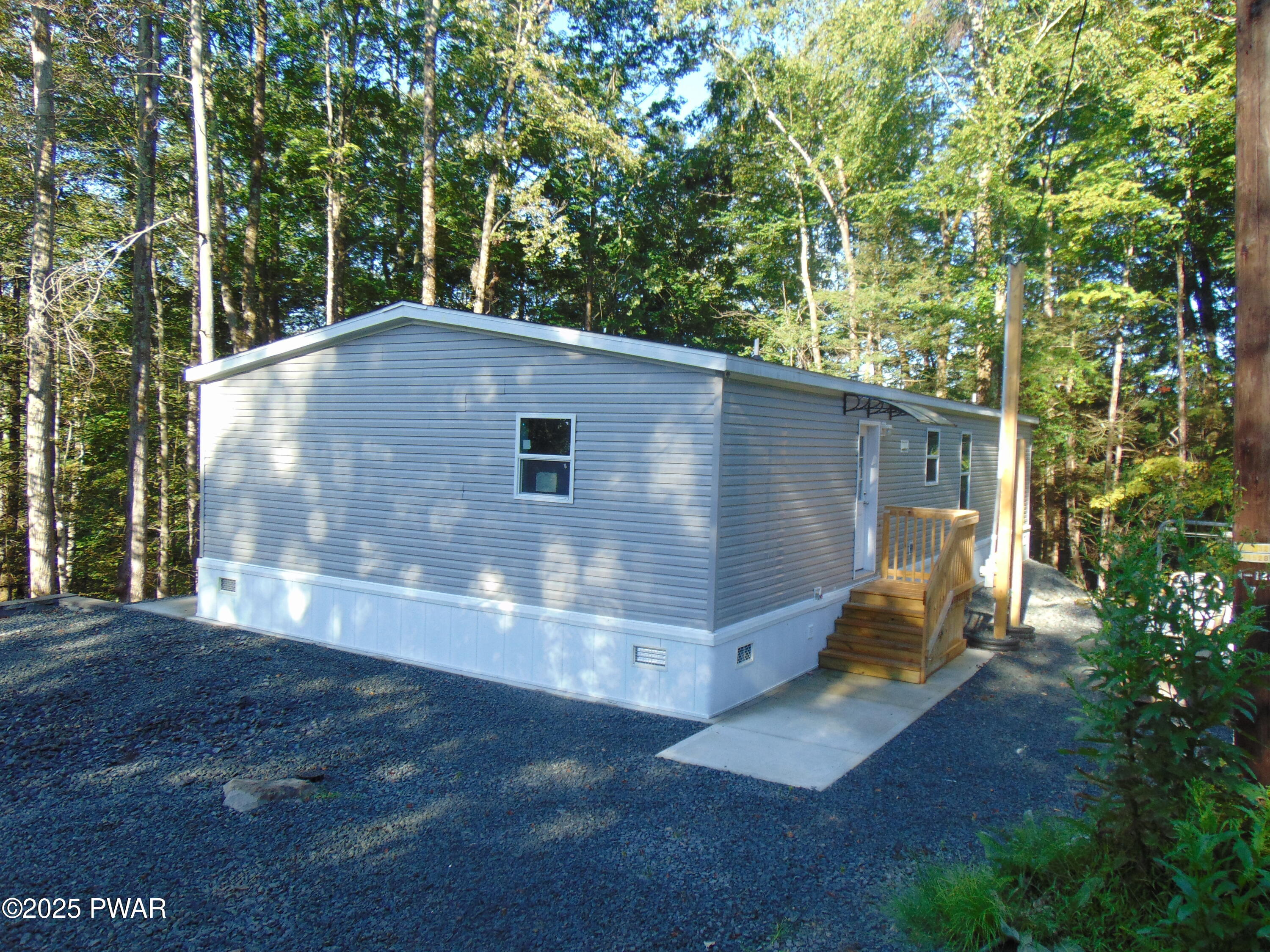 A small single-story manufactured home with gray siding sits among tall trees, with a gravel driveway and a small wooden entry stoop.
