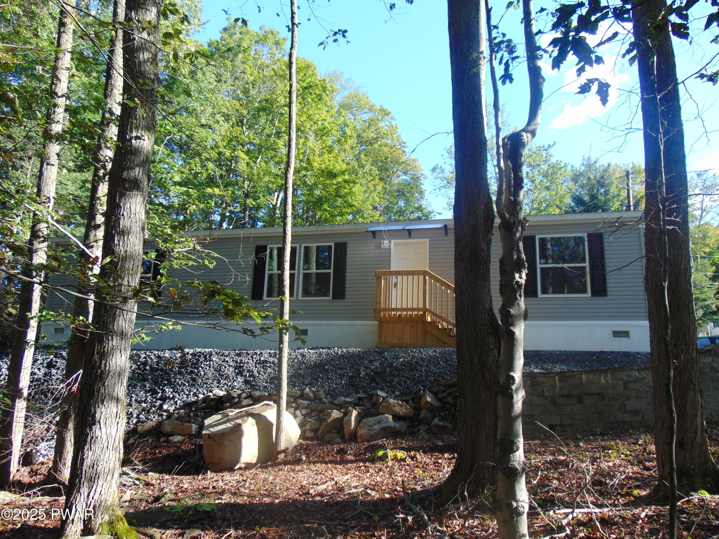 Single-story manufactured home seen from the front through trees, with gray siding, white skirting, black shutters, a small front stoop, and wooded landscaping.