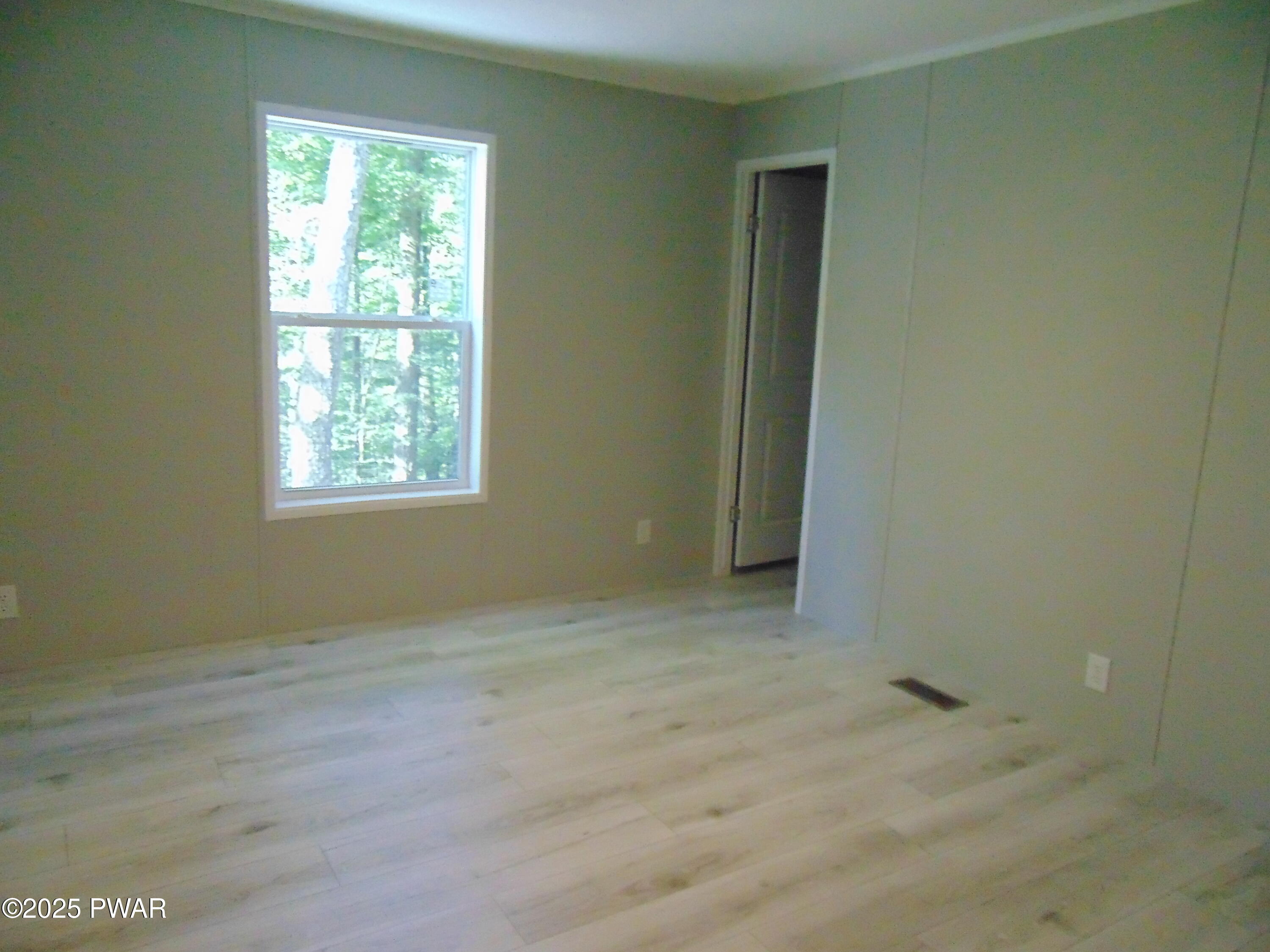 Empty bedroom with light wood flooring, gray walls, a window overlooking trees, and a closet opening.