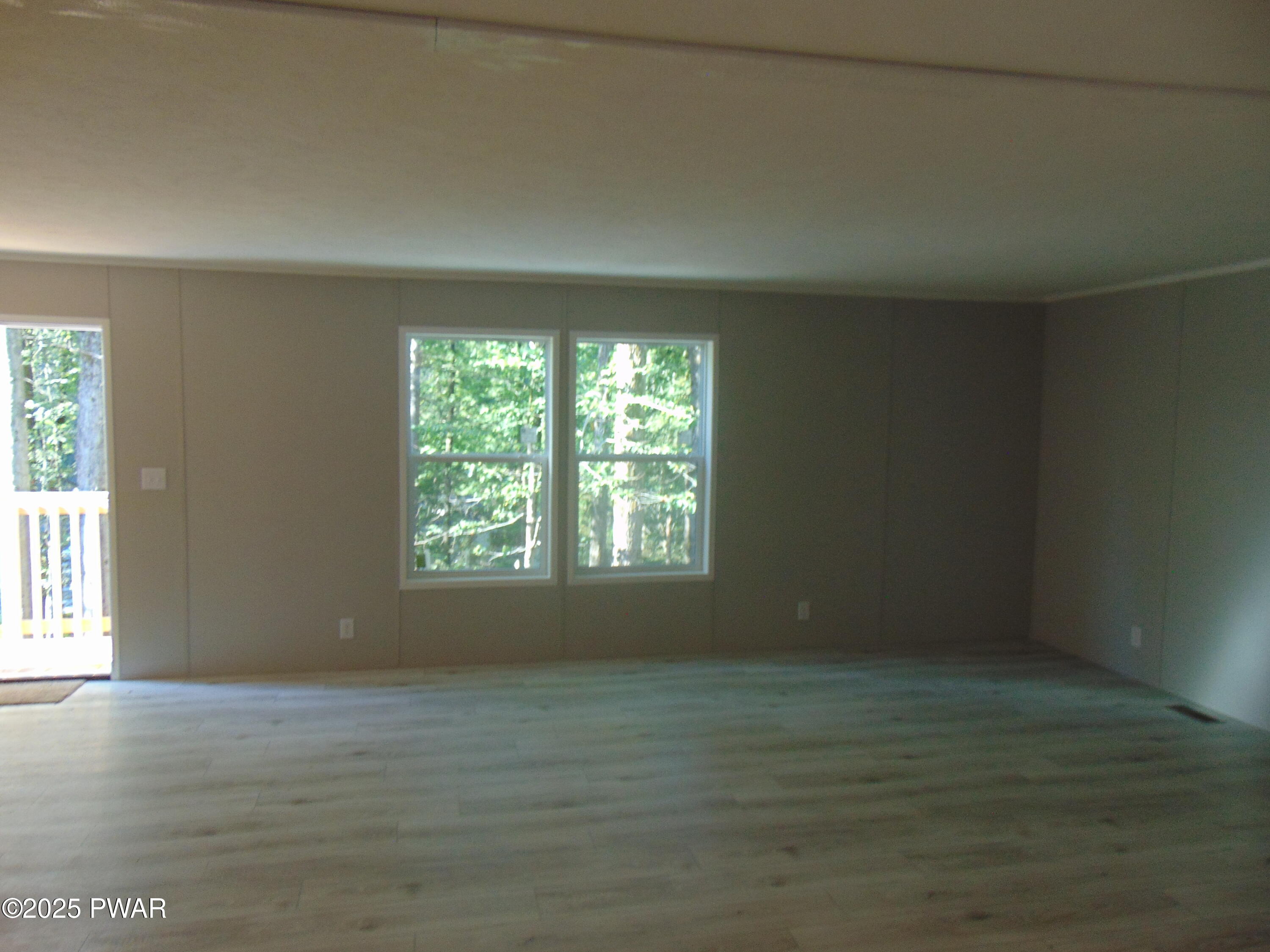 Empty living room with wood floors, beige walls, multiple windows, and a door leading outside.