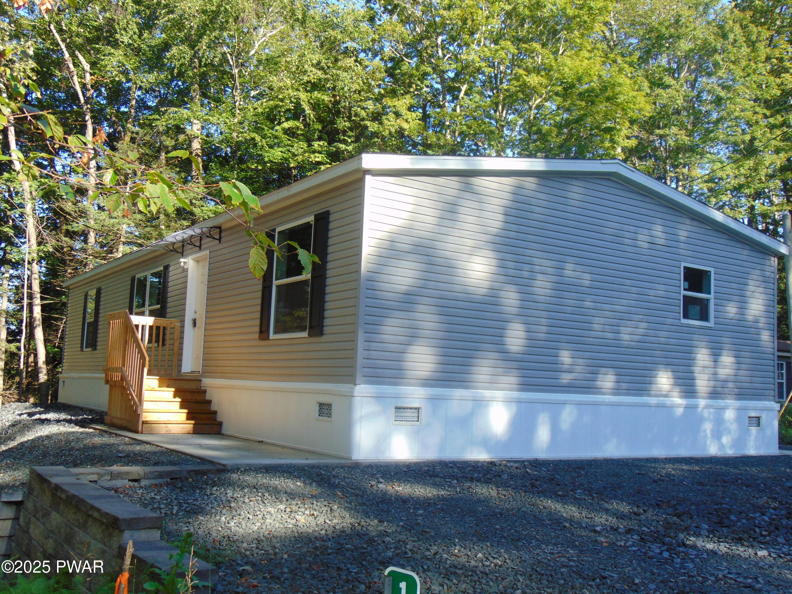 Manufactured home exterior with beige vinyl siding, small front steps and deck railing, set in a wooded lot with gravel driveway.