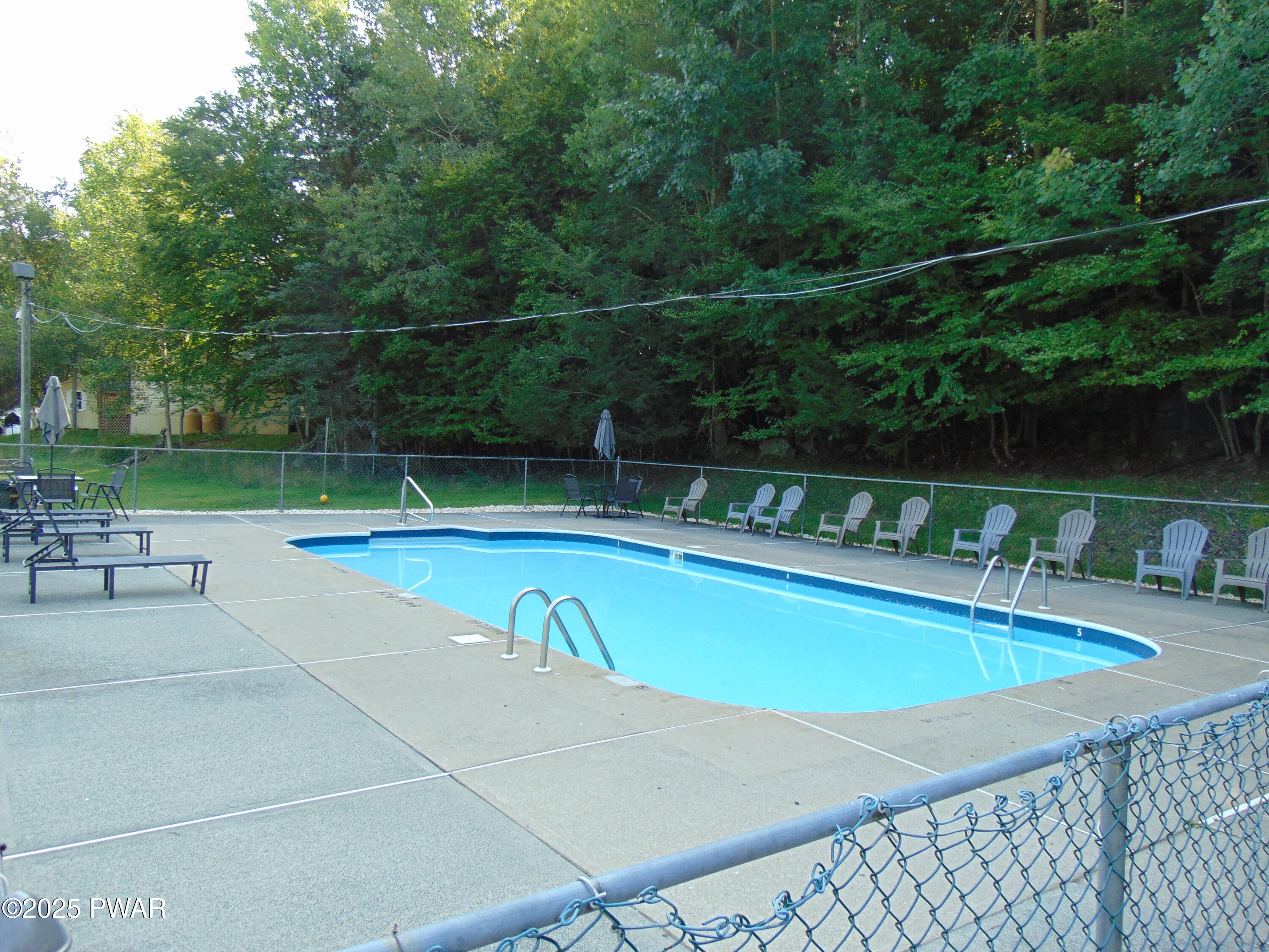 Outdoor swimming pool with concrete deck, lounge chairs, patio tables, and wooded backdrop behind a chain-link fence.
