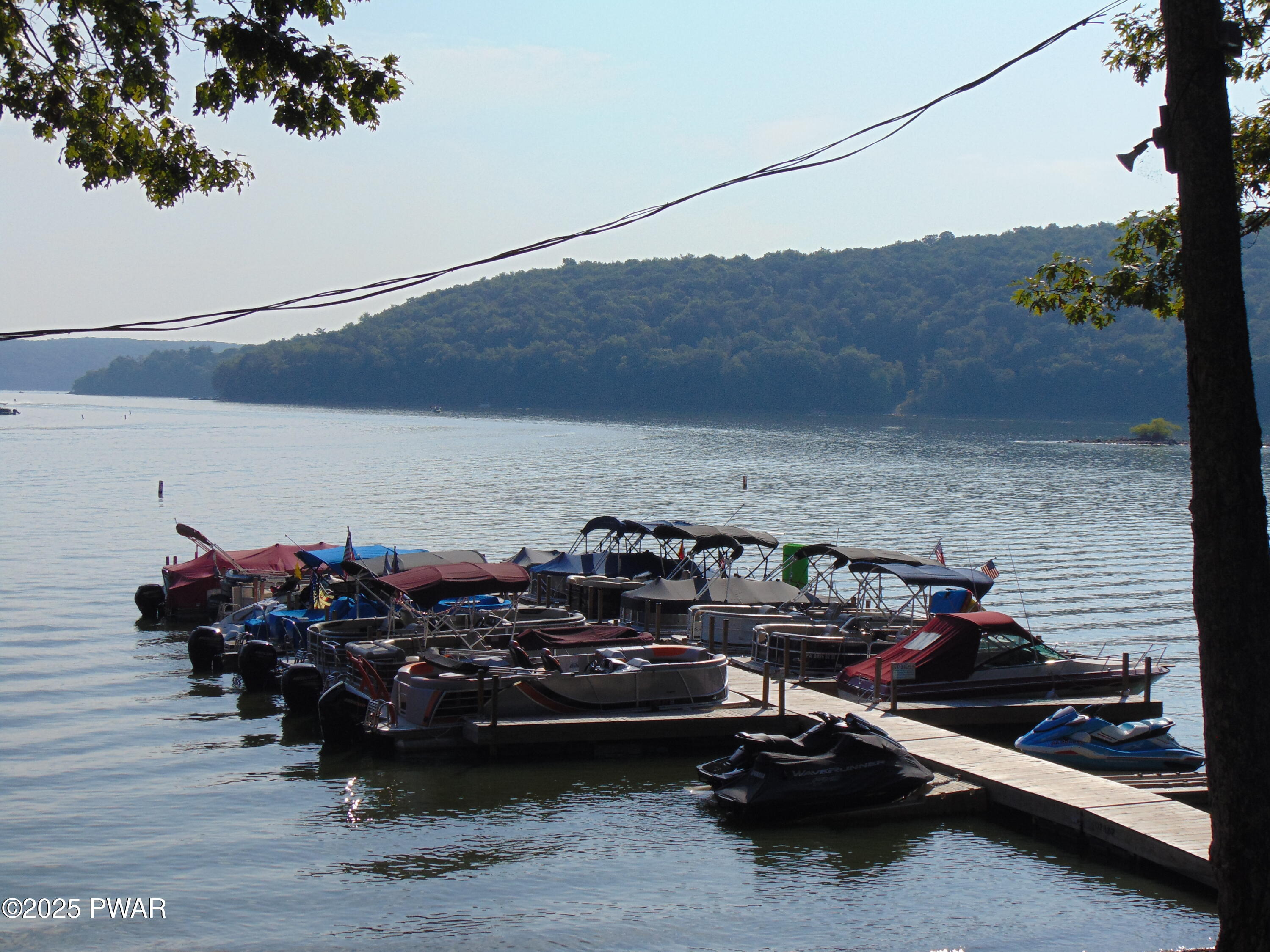 Dock with several covered boats and jet skis on a calm lake with wooded hills in the background.