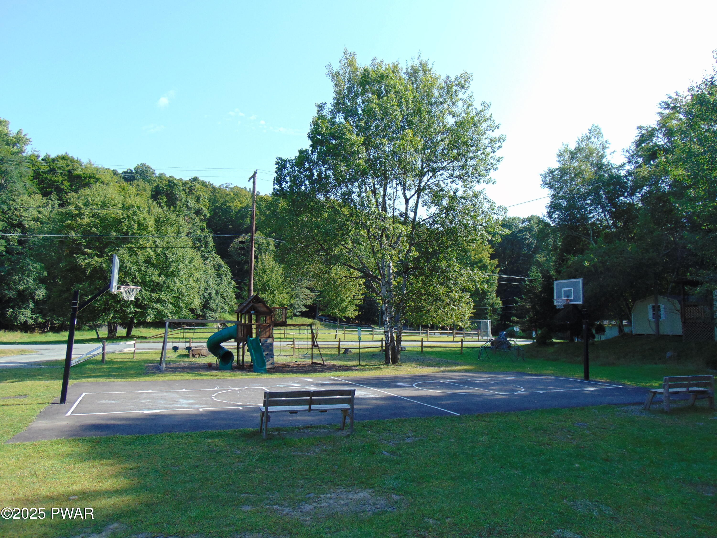 A large grassy yard with a basketball court, playground equipment, trees, and wooded surroundings.