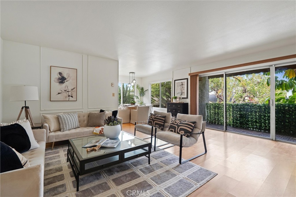 Bright living room with beige seating, a glass coffee table and patterned rug, opening through sliding glass doors to a leafy patio.