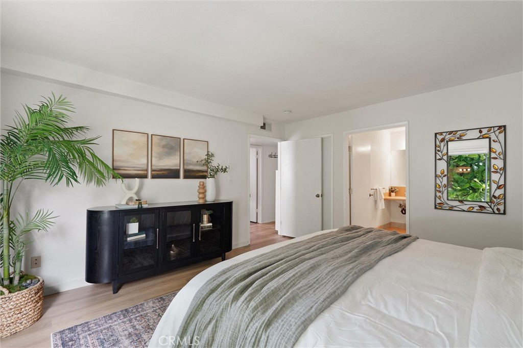 Bright bedroom at 372 Carroll E 201 with a white bed, gray-green throw, black dresser, potted palms, and an open doorway to the bathroom.