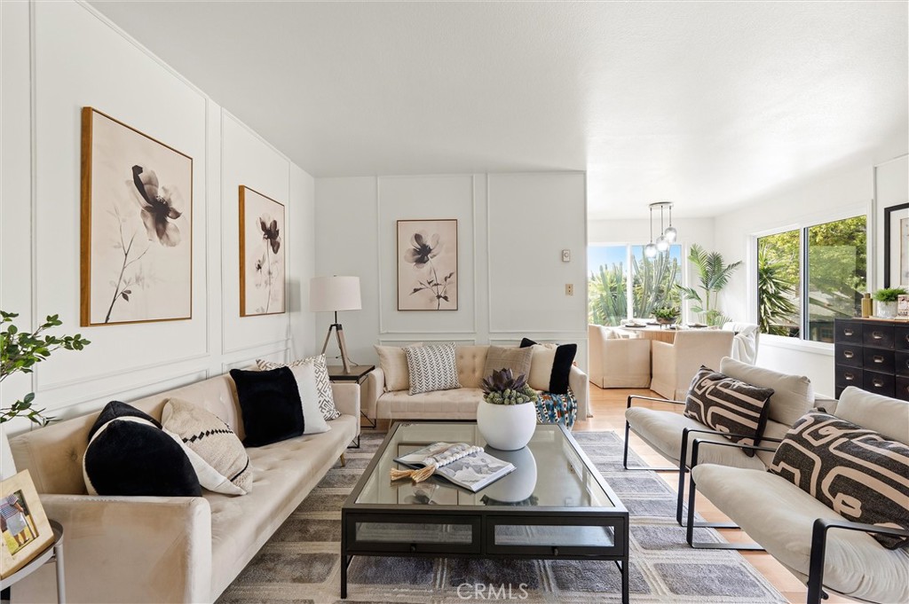 Bright living room at 372 Carroll E 201 with cream sofas, a glass coffee table, framed floral art, and large windows overlooking greenery.