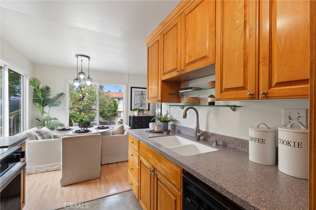 Open kitchen with warm wood cabinets, granite counters, a double sink, and stainless appliances, overlooking a bright living area with large windows.