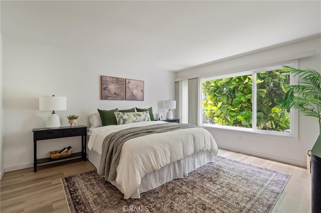 Bright bedroom at 372 Carroll E 201 with a large bed, bedside tables, hardwood floors, a patterned rug, and wide windows overlooking leafy greenery.