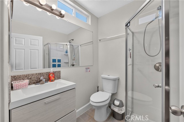 A modern bathroom featuring a white vanity with granite countertop, a toilet, and a glass-enclosed shower stall with a large mirror.