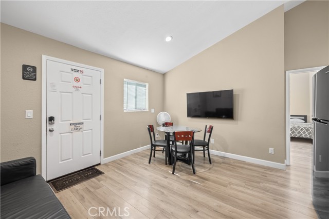 A dining area with light wood floors, a small glass round table with four black chairs, a white entry door, and a wall-mounted television, with a peek into a bedroom.