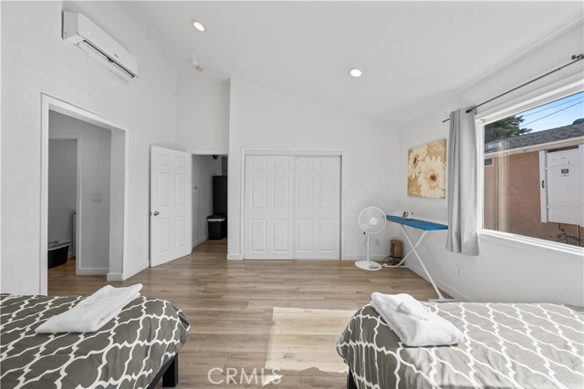 A bedroom with white walls, laminate wood flooring, two beds with gray patterned comforters, an ironing board, and a white vaulted ceiling.