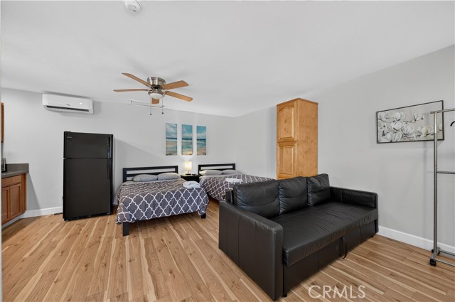 A studio apartment layout with a black leather couch in the foreground, two beds with patterned bedding, a black refrigerator, and wood flooring.