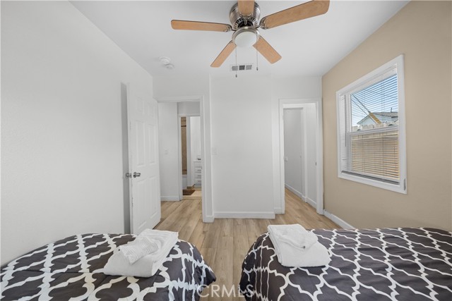 A bedroom with two beds, light wood flooring, a ceiling fan, and a view into a hallway and a bathroom.