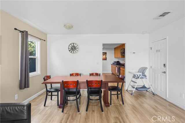 A furnished dining room with a wooden table, four chairs, and a baby high chair, featuring light wood floors and white walls with a view into the kitchen area.