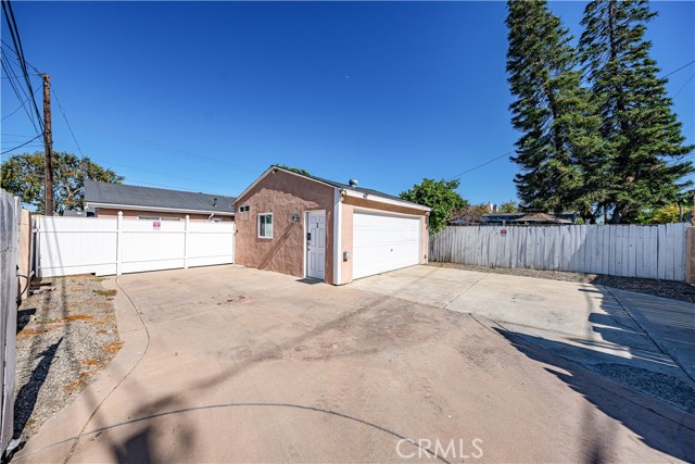 A detached single-car garage with a white roll-up door and a small side entrance, set in a spacious concrete driveway surrounded by a white picket fence under a clear blue sky.