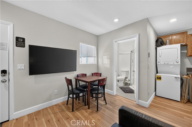A living room interior featuring a wall-mounted flat-screen television, a small dining table with four chairs, and a stacked washer and dryer unit, with a glimpse of a bathroom in the background.