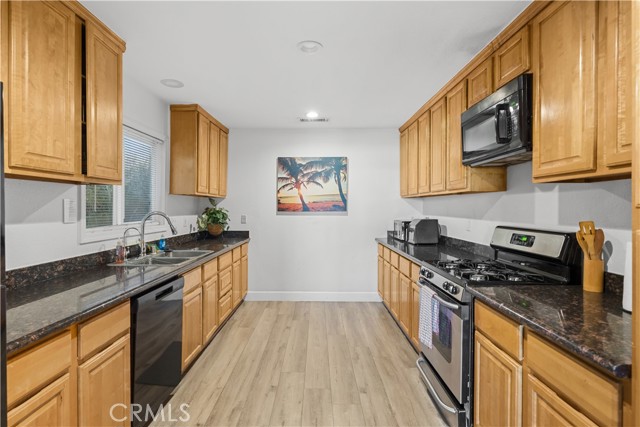 A galley-style kitchen with natural wood cabinets, dark granite countertops, stainless steel appliances, and wood-look laminate flooring.