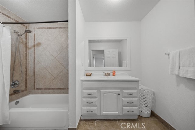 A standard bathroom with a white vanity cabinet, rectangular mirror, and a tub-shower enclosure with beige tiled walls.