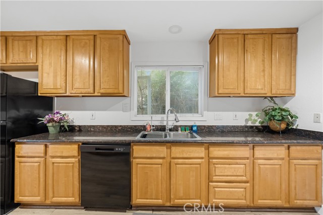 A standard kitchen featuring light wood cabinets, dark countertops, a double-basin sink, and a black dishwasher.